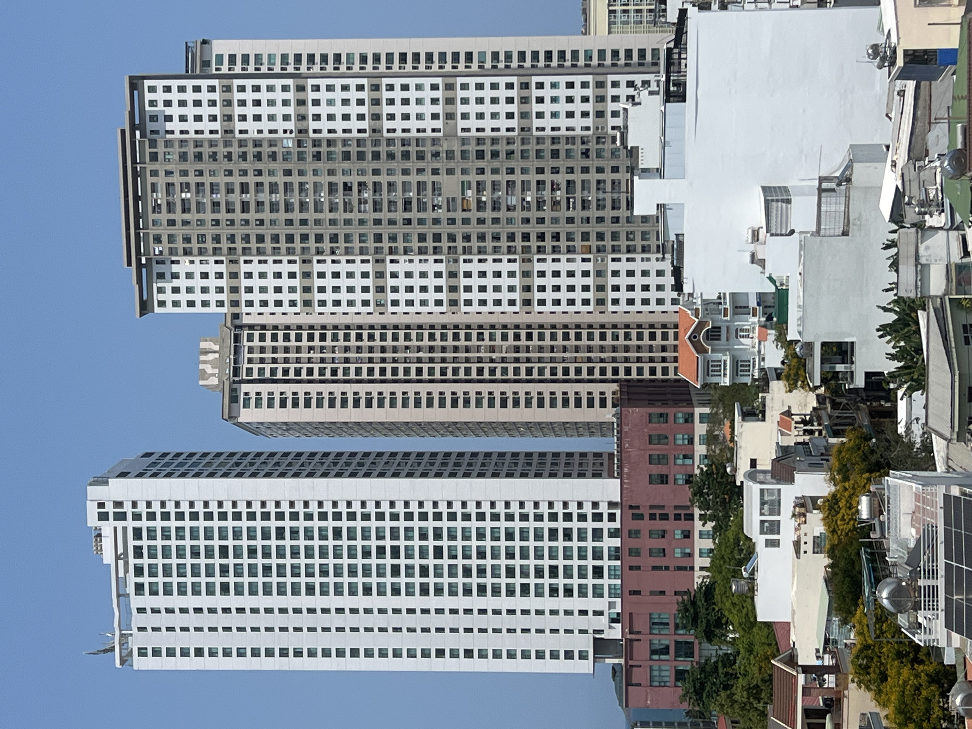 The image captures a daytime urban scene in Nha Trang, Vietnam, showcasing a mix of high-rise apartment buildings and smaller residential structures under a clear, blue sky. The buildings are characterized by repetitive window patterns, with the high-rises displaying a grid-like facade. There are other structures varying in height and architectural style, including white buildings with flat roofs, trees and other foliage visible in between the structures. The overall impression is a dense urban environment with a range of building types, reflecting the city's landscape.
