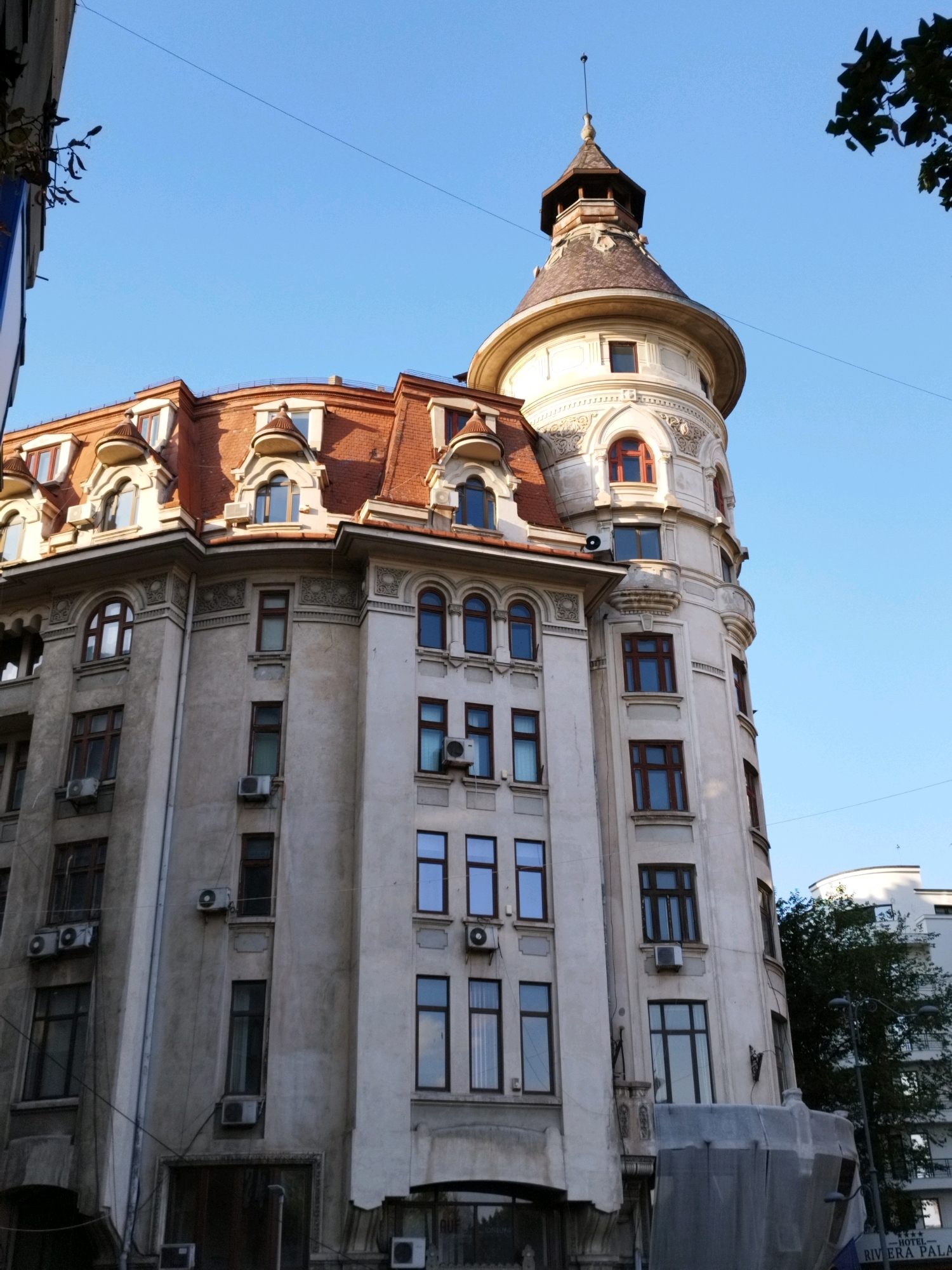This image shows a detail of the Hotel Rivoli building, located at Calea Victoriei 222, Bucharest, Romania. The building features a distinctive turret with a pointed roof, reminiscent of Art Nouveau architecture. The facade is a light color, contrasting with the red-tiled roof and dark-framed windows. Several windows have air conditioning units installed. The sky is clear and blue, with some tree branches visible in the upper right corner.