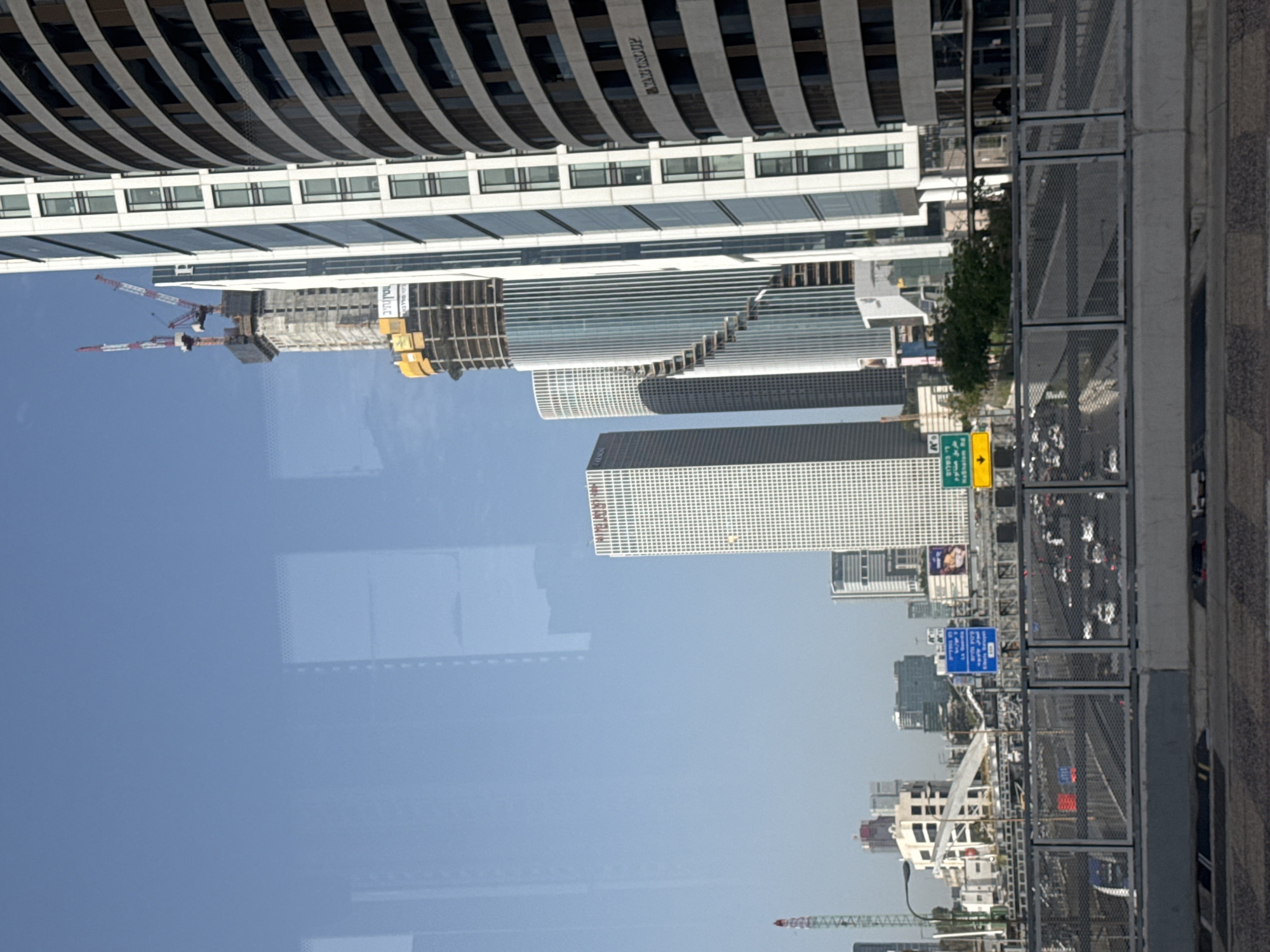 The image depicts a cityscape in Tel Aviv, Israel. The shot appears to be taken from an elevated position, possibly a building or overpass, overlooking a busy highway with vehicular traffic. Several high-rise buildings and skyscrapers dominate the skyline, including some under construction with cranes. The sky is mostly clear, with a slight haze. The image is rotated 90 degrees counterclockwise.
