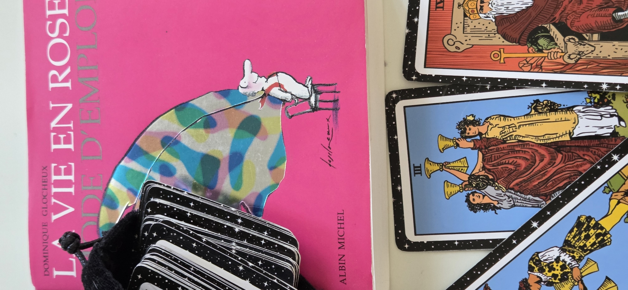 The image shows a close-up view of a book and a stack of tarot cards. The book, titled "LA VIE EN ROSE MODE D'EMPLOI" by Dominique Glocheux and published by Albin Michel, has a bright pink cover. A small, black velvet pouch, likely containing a tarot deck, is partially visible on the left side of the book. A whimsical illustration of a person sitting on a chair with a colorful, translucent sheet behind them is also on the book cover. Below the book, several tarot cards are laid out. The visible cards include the Three of Cups, featuring three women holding cups, and a portion of a card depicting a crowned figure seated on a throne, possibly the Emperor. The tarot cards have black borders with white stars. The scene is well-lit, and the focus is sharp on the book and the tarot cards. There is no discernible information in the image to indicate its location in the city of Strasbourg, France.