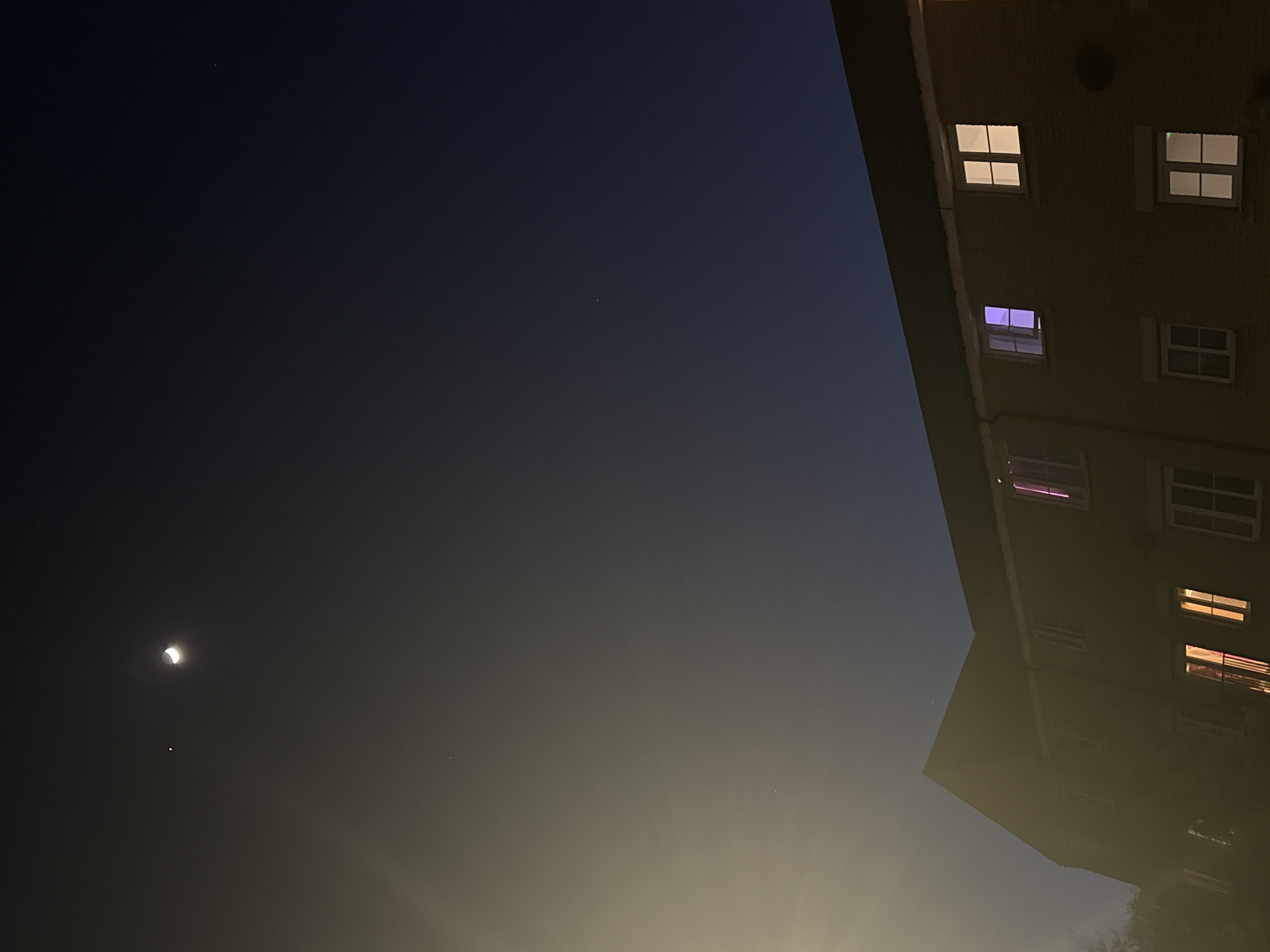 The image captures a night scene looking upwards at a crescent moon and a portion of a building in Great Yarmouth, United Kingdom. The sky occupies the majority of the frame, transitioning from a dark, deep blue at the top to a luminous yellow-orange gradient towards the bottom right, indicative of light pollution from an urban environment.
A prominent, bright crescent moon is visible in the upper-left quadrant, its illuminated edge facing right. Below it, a notably bright star or planet can be seen, accompanied by several other faint stars scattered across the dark expanse of the sky. The absence of clouds suggests clear weather conditions.
On the right side, a multi-story brick building is partially visible, extending from the top to the bottom of the frame. It appears to be an apartment block or similar structure. Numerous windows adorn its facade, some of which are illuminated from within. Several windows glow with white light, while others display distinct colours: one emits a bright purple light, another a softer purple/pink, and a lower one glows orange/pink. The windows feature white frames, with some showcasing internal grid patterns.
The scene conveys a tranquil, nocturnal atmosphere in an urban or suburban setting. No specific activities or people are depicted. There is no visible text in the image.