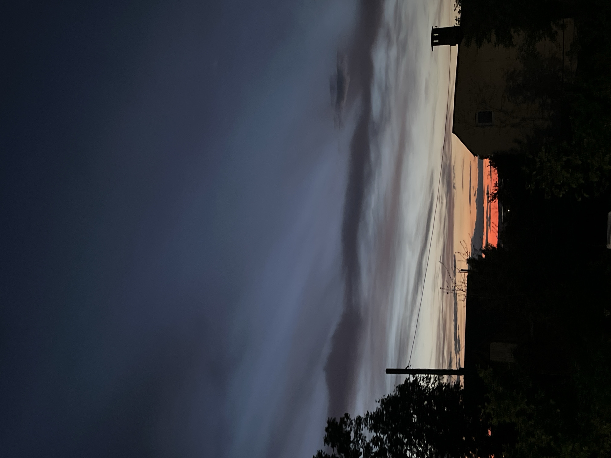 The image shows a building and a cloudscape, seemingly at dusk or dawn. The left side is dominated by a dark blue sky, possibly due to heavy cloud cover or nightfall. Toward the right, there are striations of light and color in the sky, indicating a sunset or sunrise. The presence of a utility pole suggests a residential or suburban area. A building with a chimney is on the right side of the image, partially obscured by trees. The location is possibly Chaikovsky, Russia, based on the prompt.