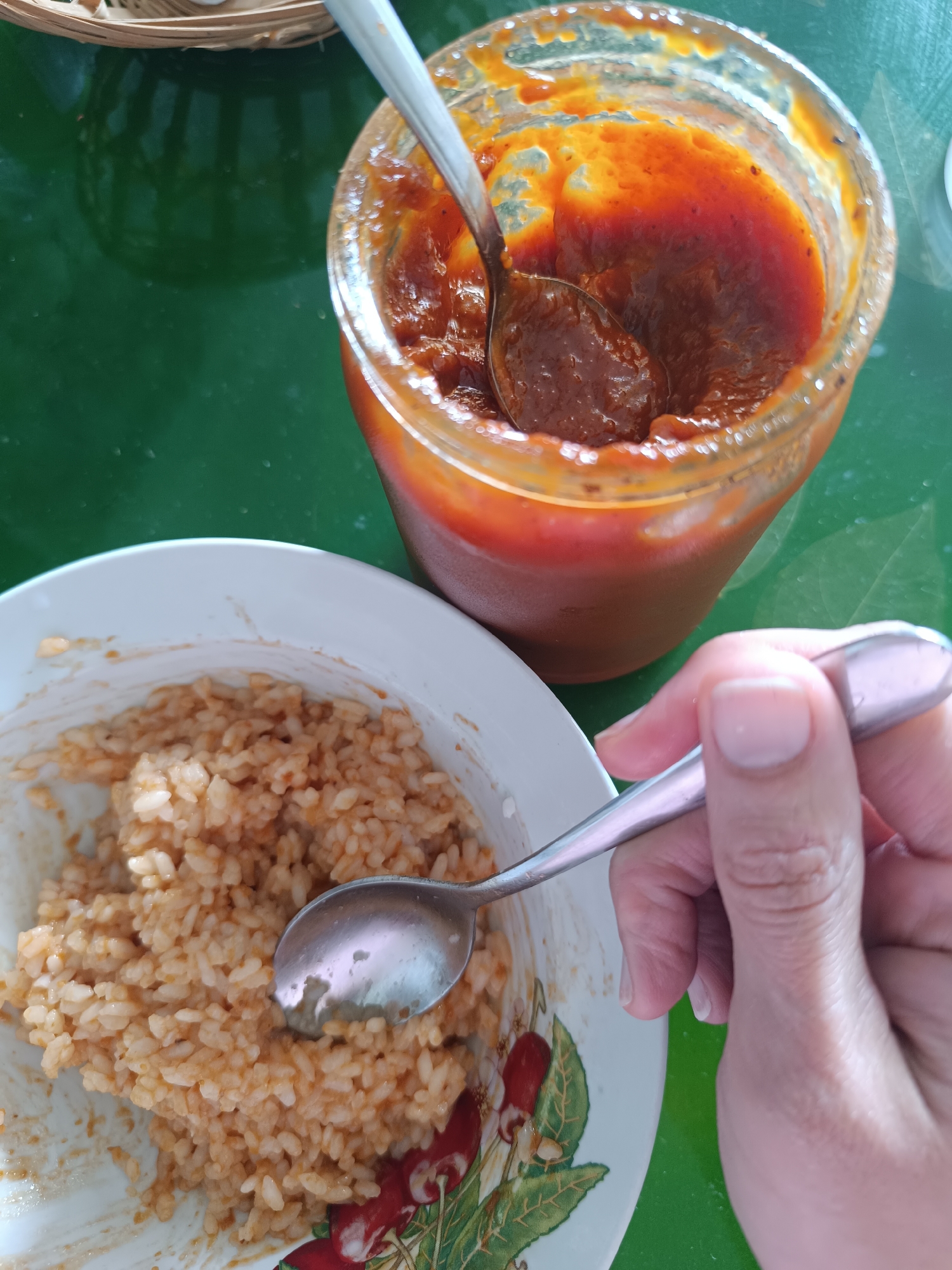 On a green surface, a bowl of rice and a jar of red sauce are visible. A hand holds a spoon over the rice. The bowl is white with a cherry design around the edge. The jar appears to be glass, and a spoon is inside the jar of red sauce. The scene appears to be a meal setting. The location is Lermontov, Russia.