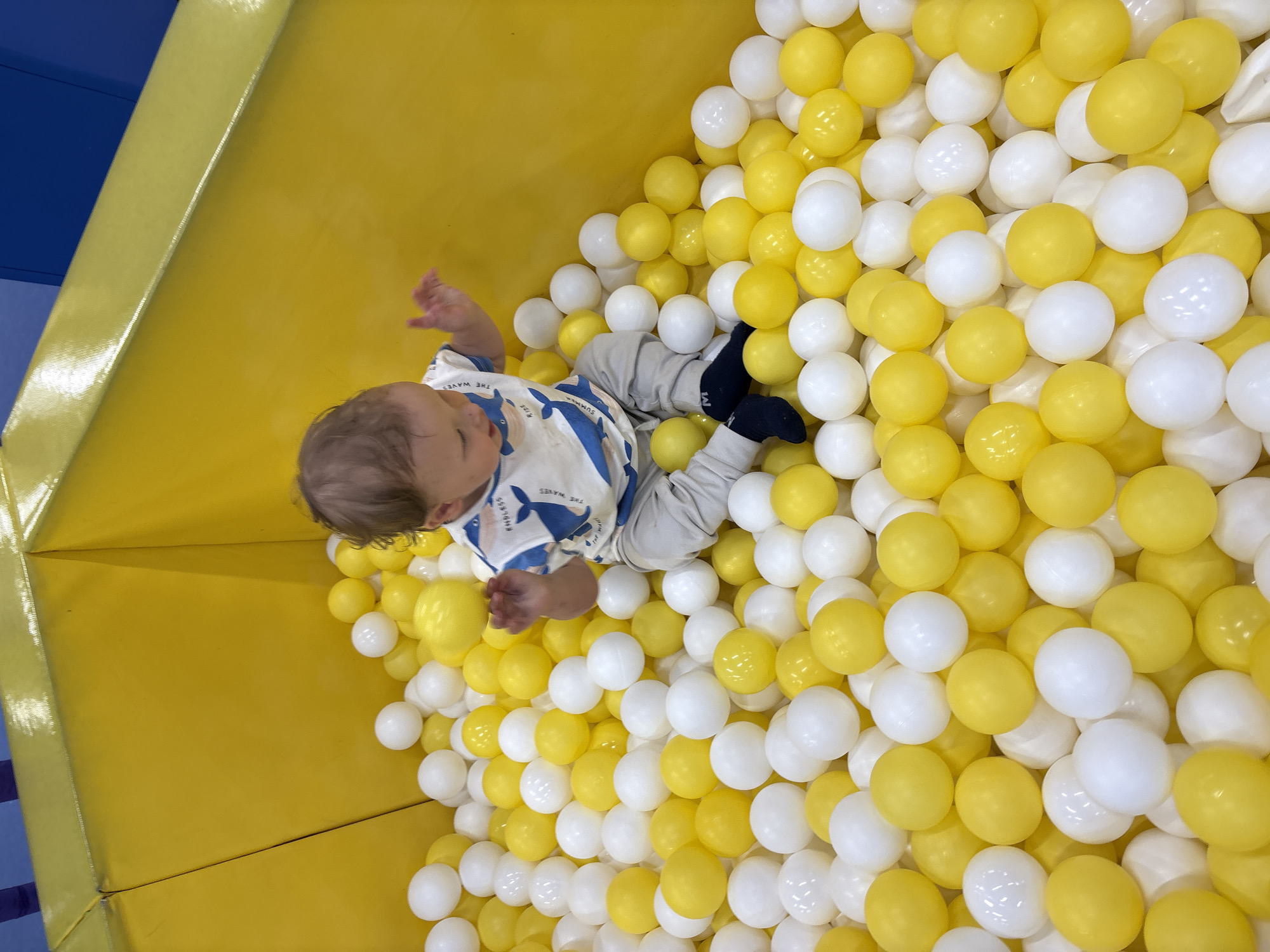 A young child sits in a ball pit filled with yellow and white plastic balls. The ball pit is surrounded by a yellow padded wall with a blue cushioned edge visible in the upper-left corner. The child is wearing a patterned shirt and gray pants with dark socks. The location is in Izhevsk, Russia.