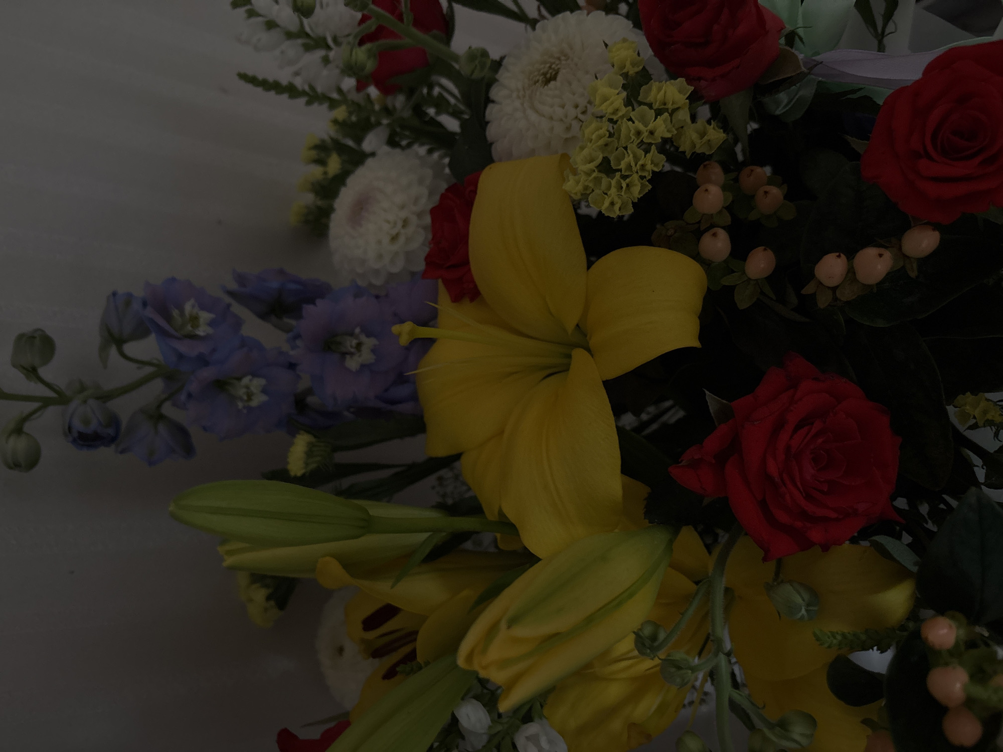 The image displays a close-up, overhead view of a vibrant floral arrangement. The arrangement features several types of flowers in various colors, including bright yellow lilies, deep red roses, and clusters of small white and pale blue flowers, possibly delphiniums. There are also small, light orange berries interspersed among the foliage. The background is a plain, light-colored wall. The lighting is dim, suggesting an indoor setting. The image does not provide any information about the location in the city of Shepetivka, Ukraine, nor does it depict any people or specific actions.