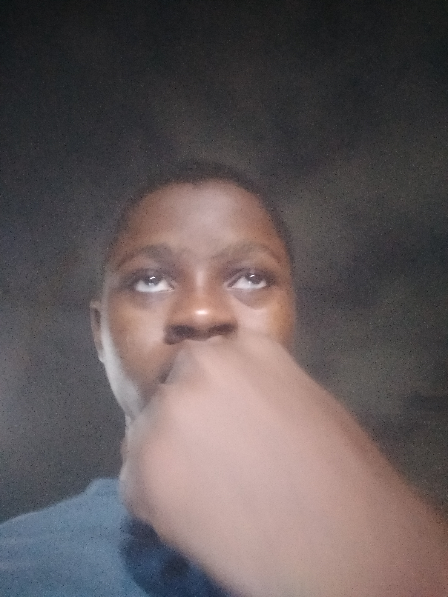 The image depicts a person looking upwards, with a hand partially obscuring their face. The person has dark skin, short hair, and is wearing a dark blue shirt. The background is a dark, nondescript wall. The location, Shangisha, Nigeria, cannot be verified from the image.