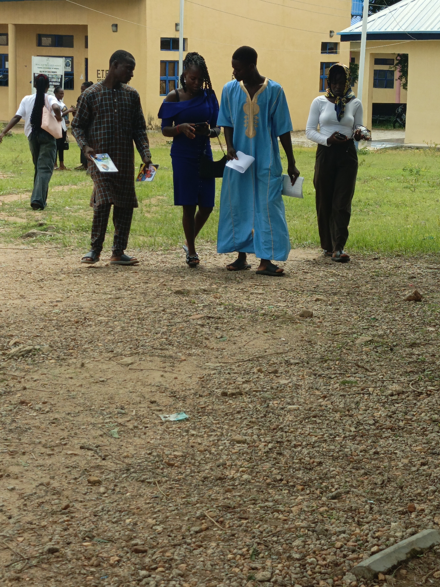 A group of five individuals are walking on a gravel path in front of a yellow, two-story building. The building has blue-trimmed windows and appears to be a school or administrative office. On the left, a person wearing a white t-shirt and dark pants walks away from the camera, carrying a light pink bag. Next to them, a man in a plaid tunic and pants walks towards the camera, holding two open books. To his right, a woman in a blue off-the-shoulder dress is looking down at her phone. Further to the right, a man in a blue robe with gold trim walks towards the camera, holding a piece of paper. Beside him, a woman wearing a white long-sleeved shirt and dark pants, with a patterned headscarf, is looking down at her phone. The environment is outdoors, with green grass behind the building and on the right side of the path. The ground is covered in small stones and dirt.