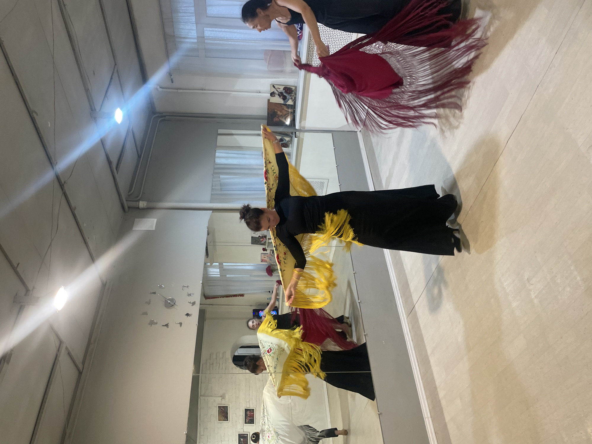 In Sochi, Russia, within a dance studio featuring a large mirrored wall, three women are engaged in flamenco dancing. The woman in the foreground, wearing a black dress, is holding a yellow shawl with fringe, her arms outstretched. To her right, another woman in a black tank top and pants is twirling a dark red shawl with extensive fringe. Further back, a third woman in a black dress is partially obscured, holding a yellow shawl similar to the foreground dancer's, and a red skirt is visible around her legs. The studio has light-colored wooden flooring and a gray ceiling with exposed conduits and fluorescent lighting. A wall-mounted clock with Roman numerals is visible on the wall. The mirrored wall reflects the dancers and the studio's interior, including several framed artworks on the wall. The overall scene captures a moment of dynamic movement and performance.