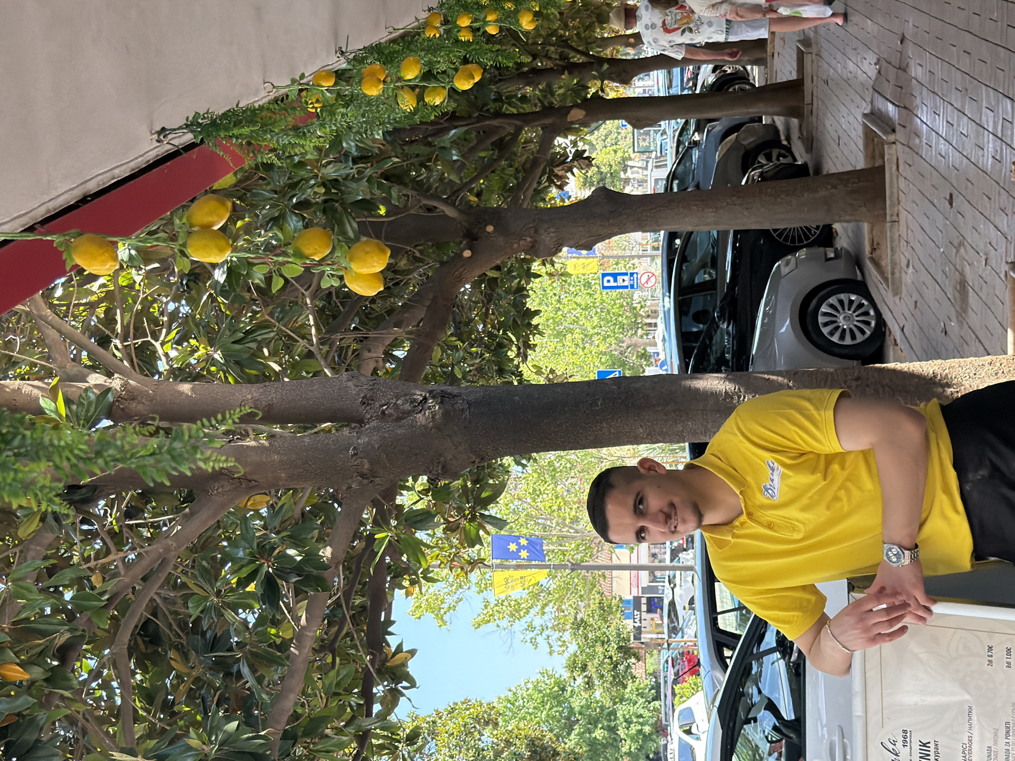 In Budva, Montenegro, a young man wearing a bright yellow polo shirt and black shorts smiles at the camera while standing next to a car. He has a silver watch on his left wrist and a bracelet. The scene appears to be outdoors, with trees laden with lemons providing shade. In the background, parked cars line a street, and buildings suggest an urban environment. The sky is visible above, indicating it's daytime, likely a sunny day. Two flags, one blue and one yellow, are flying on poles in the middle ground. Some text is visible on the side of the car, including "Branka," "1968," and "NIK," along with pricing in Euros for beverages. The overall impression is a casual, sunny day in a European town.