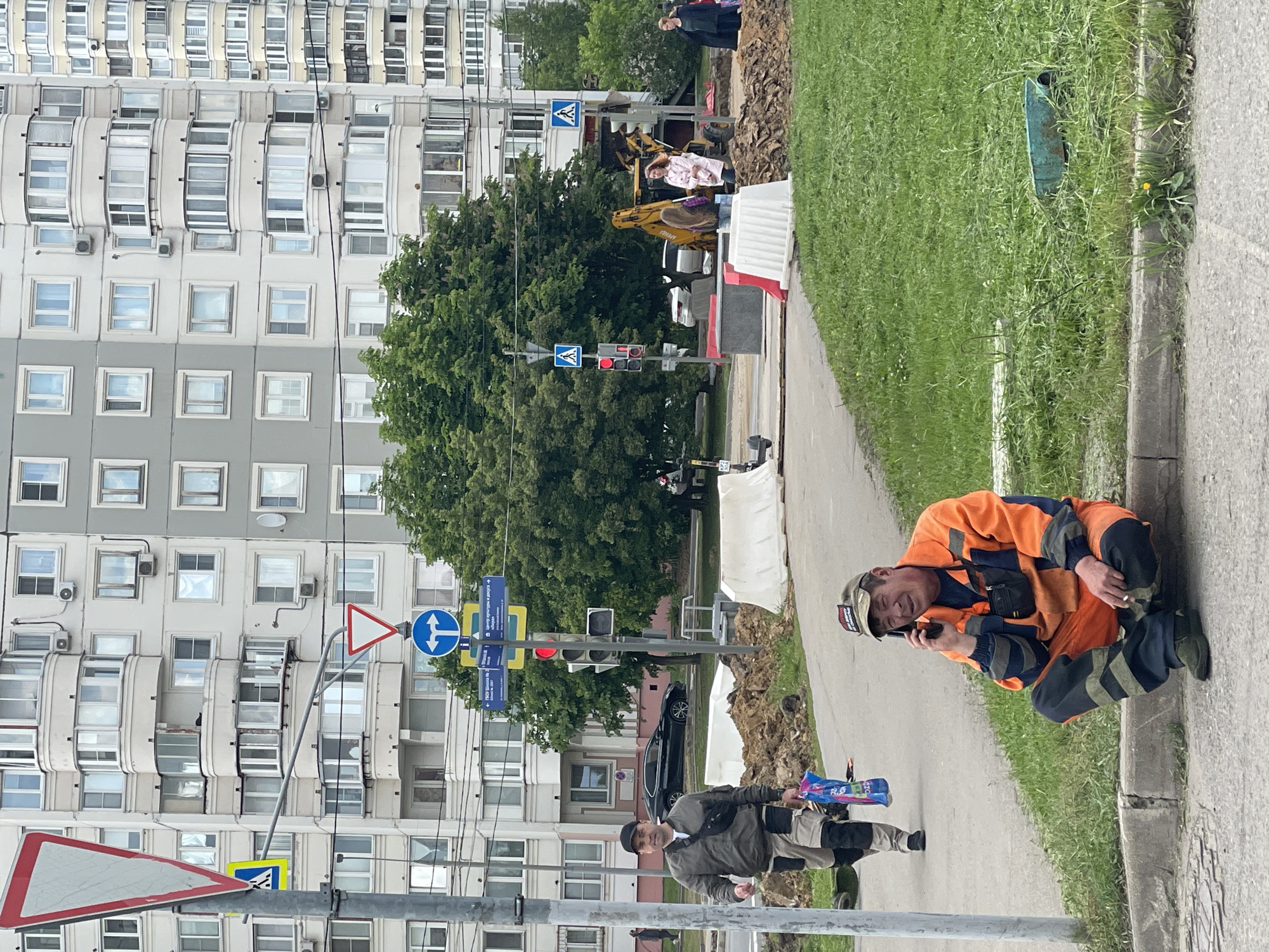 The image captures a street scene in Moscow, Russia, featuring construction and everyday life. A man in an orange work suit is kneeling on the pavement, talking on his phone. Another man is walking towards him carrying a blue package. In the background, a construction site is visible with a yellow excavator and a few people standing around. There are road signs and a traffic light indicating a crosswalk. A large building with multiple windows occupies a significant portion of the background. On the other side of the road there is green grass. A blue skateboard is leaning in the grass.