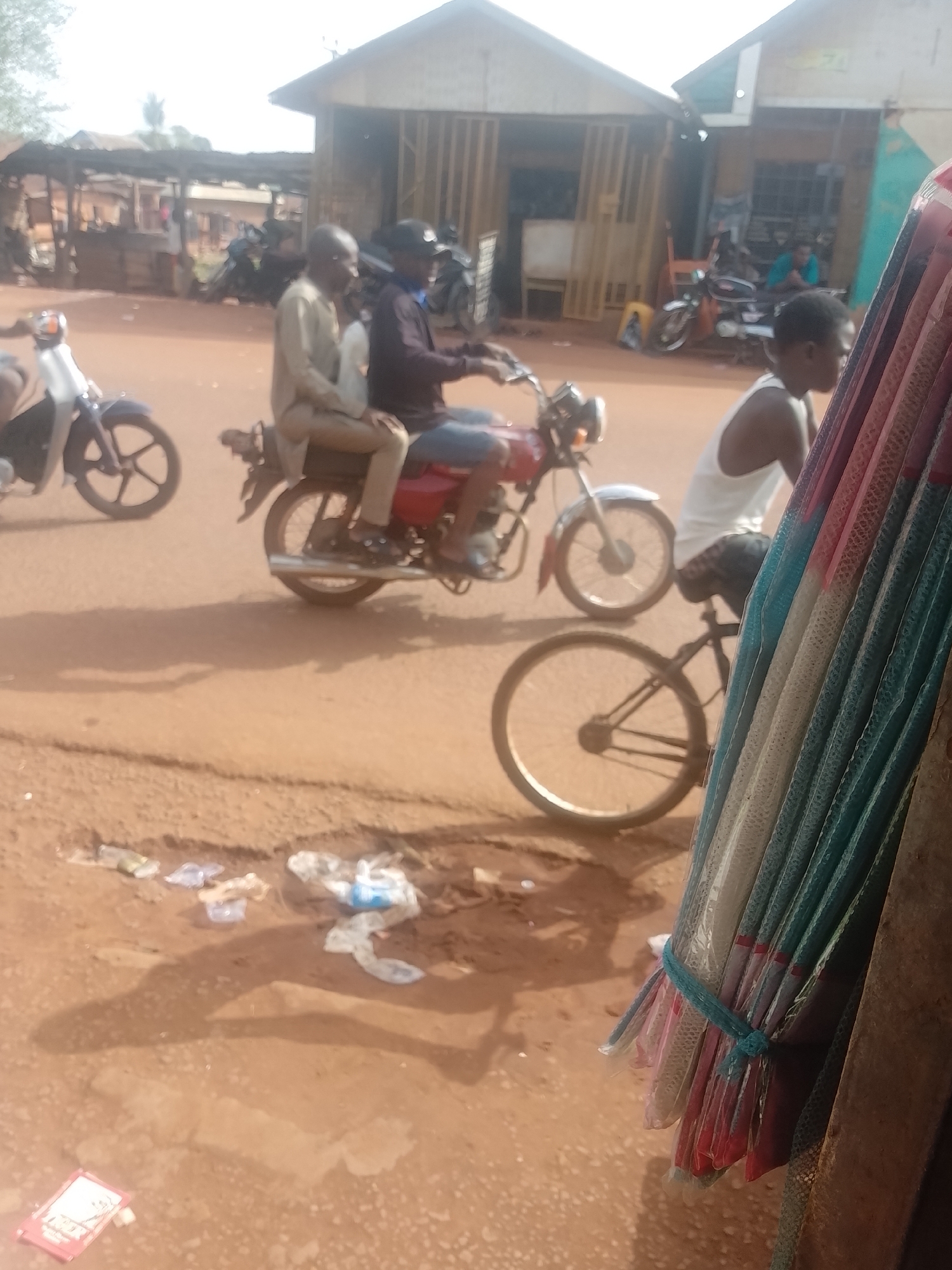 The image shows a street scene in Ejule, Nigeria. The ground is reddish-brown. Litter, including plastic bags and wrappers, is visible on the ground.
Several people are present, including a man on a bicycle wearing a white tank top, and two men riding a red motorcycle. The driver wears a dark shirt, while the passenger wears a light-colored shirt. Another motorbike is visible parked in the background.
Background elements include buildings made of wooden structures. Other motorbikes are parked in front of one of the buildings.
A bundle of items wrapped in colorful plastic is visible in the foreground, attached to the corner of a building.