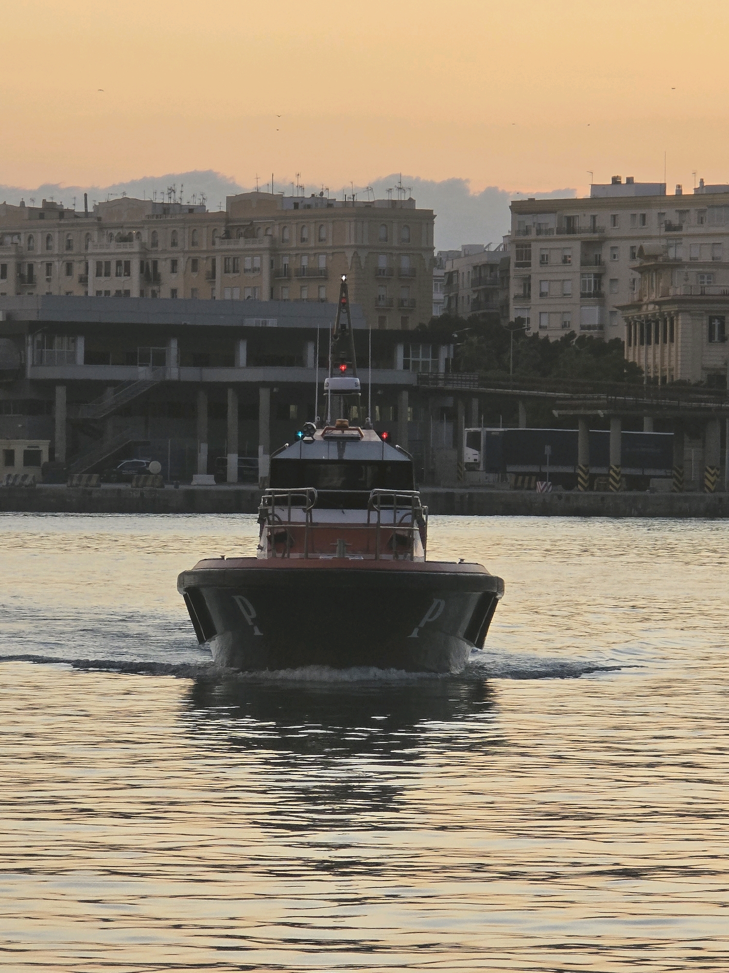 A law enforcement vessel, identified by the letters "P" on its bow, moves across calm, rippled water. The boat has a red and black hull and a white cabin with red and green lights on top. In the background, a city skyline is visible. Buildings of various heights and architectural styles line the waterfront, with some appearing to be apartment blocks or office buildings. A grey elevated walkway or bridge with concrete supports spans across the midground, and a dark-colored truck is visible beneath it. The sky is a soft orange and yellow, suggesting either sunrise or sunset, with faint clouds and distant birds. The scene appears to be the Port of Málaga, Spain, due to the distinctive architecture and port infrastructure. No people are visible on the boat or in the background.