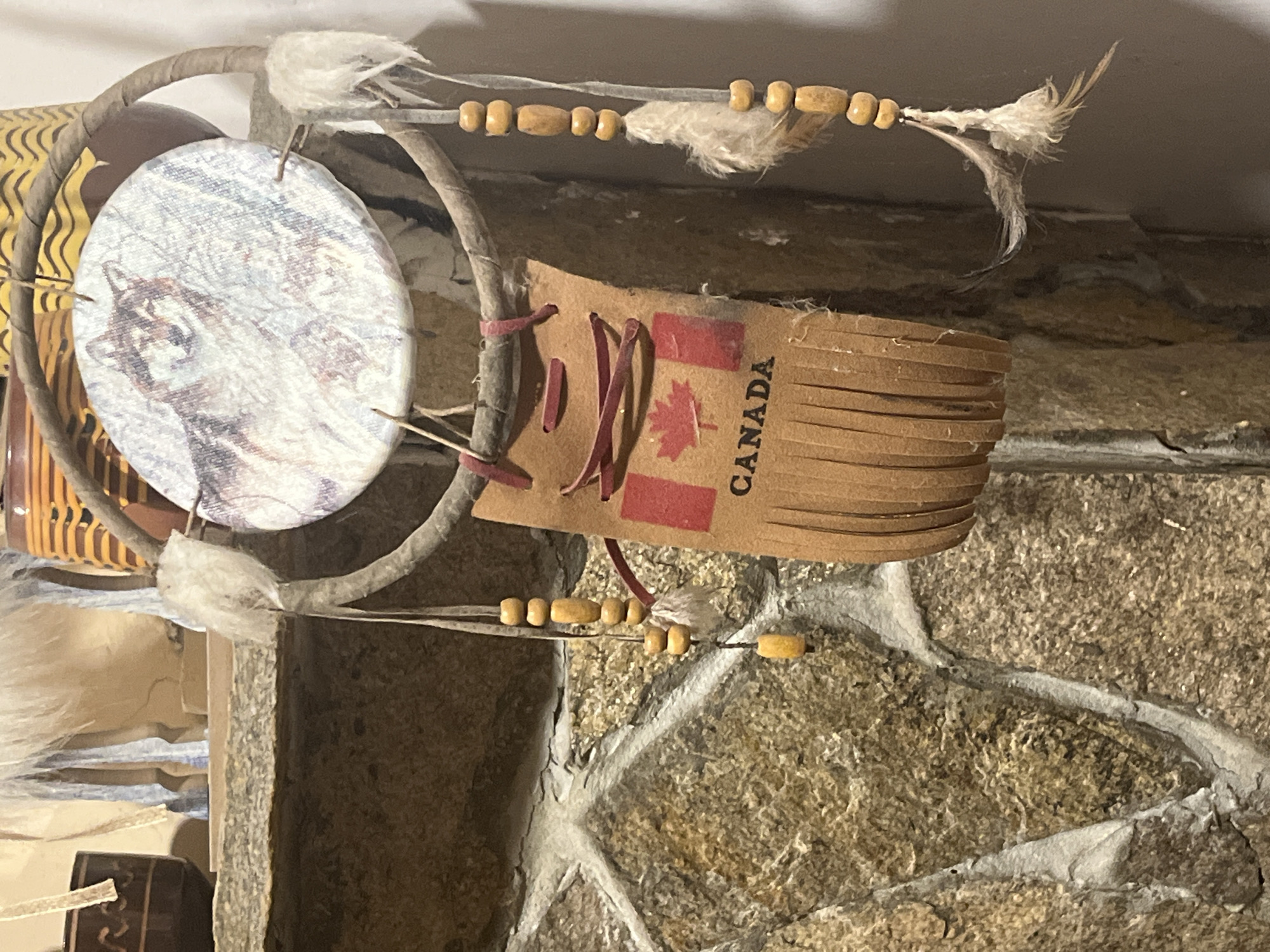 The image shows a dreamcatcher hanging against a stone wall. The dreamcatcher has a central circular disc with a wolf image, a loop made of a branch with white tufts and feathers attached, beaded strings, and a tag featuring the Canadian flag and the word "CANADA". The stone wall appears to be interior. The location in Varshilo, Bulgaria is not evident.