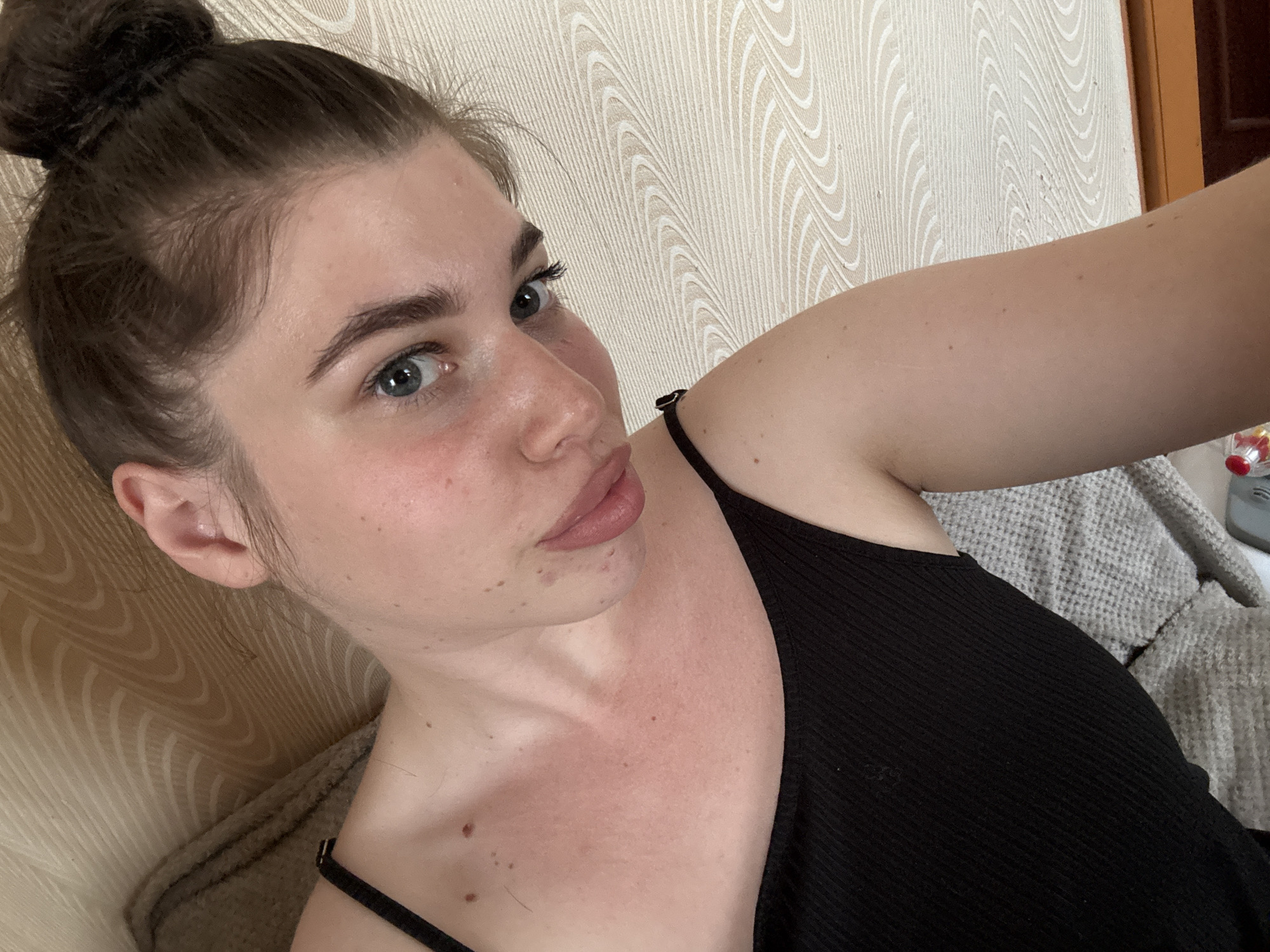 A selfie of a woman with her hair in a bun, wearing a black tank top. The background features a patterned wall and a glimpse of bedding. The location cannot be determined from the image.