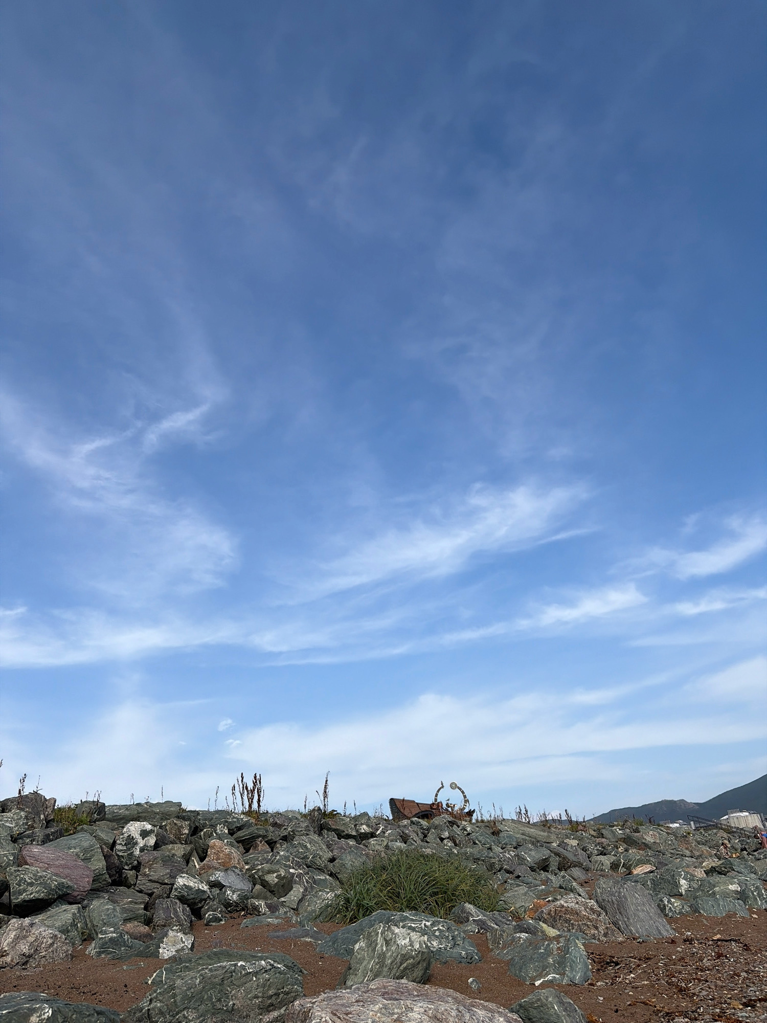 The image depicts a rocky shoreline with a clear blue sky filled with wispy white clouds. In the foreground, a jumble of large, grey and reddish-brown rocks forms a rugged embankment. Some sparse, dry vegetation and green grass are scattered amongst the rocks. In the middle ground, a unique wooden bench with an ornate, curved metal arch is visible, suggesting a public space or park. Further in the background, a hilly landscape with some buildings is discernible. A person in blue shorts is visible on the far right, near the water's edge. This scene is located in Korsakov, Russia, likely an area developed for public recreation along the coast.