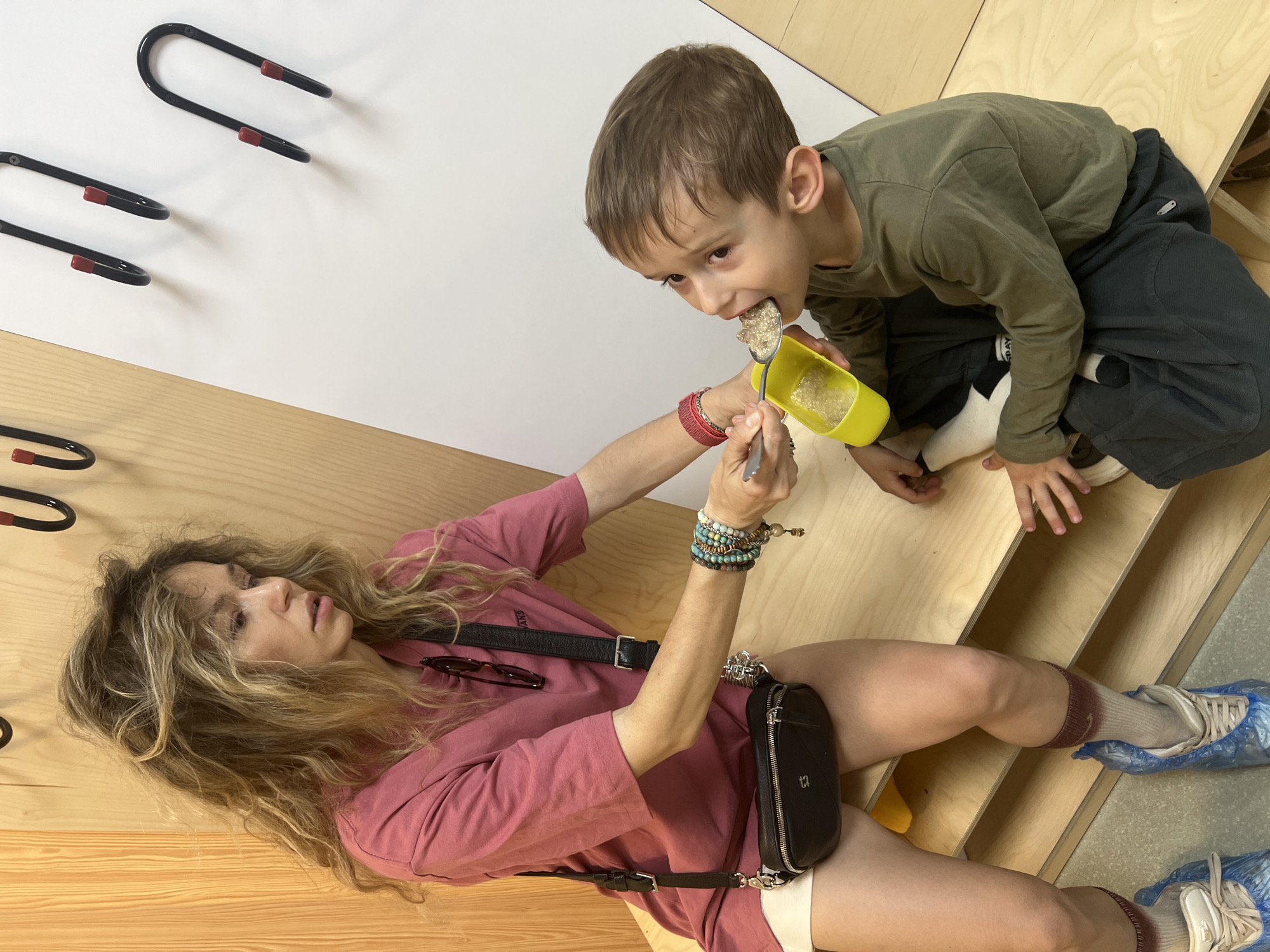 In this medium shot image, a woman with long, wavy blonde hair is lying down on a wooden surface, propping herself up with her arm. She is wearing a pink t-shirt, shorts, and a black fanny pack around her waist. She has on several bracelets and a crossbody strap from her fanny pack is visible. She is holding a spoon and a yellow cup, and appears to be feeding a young boy. The boy, wearing a green long-sleeved shirt and dark pants, is sitting up and eating from the spoon. He is looking towards the camera with his mouth open. His feet are covered in blue plastic shoe covers. The background shows a white wall with black hooks and a wooden wall and floor. The scene appears to be indoors. The lighting suggests it could be daytime. The overall mood seems casual and familial, with the act of feeding indicating care and interaction.