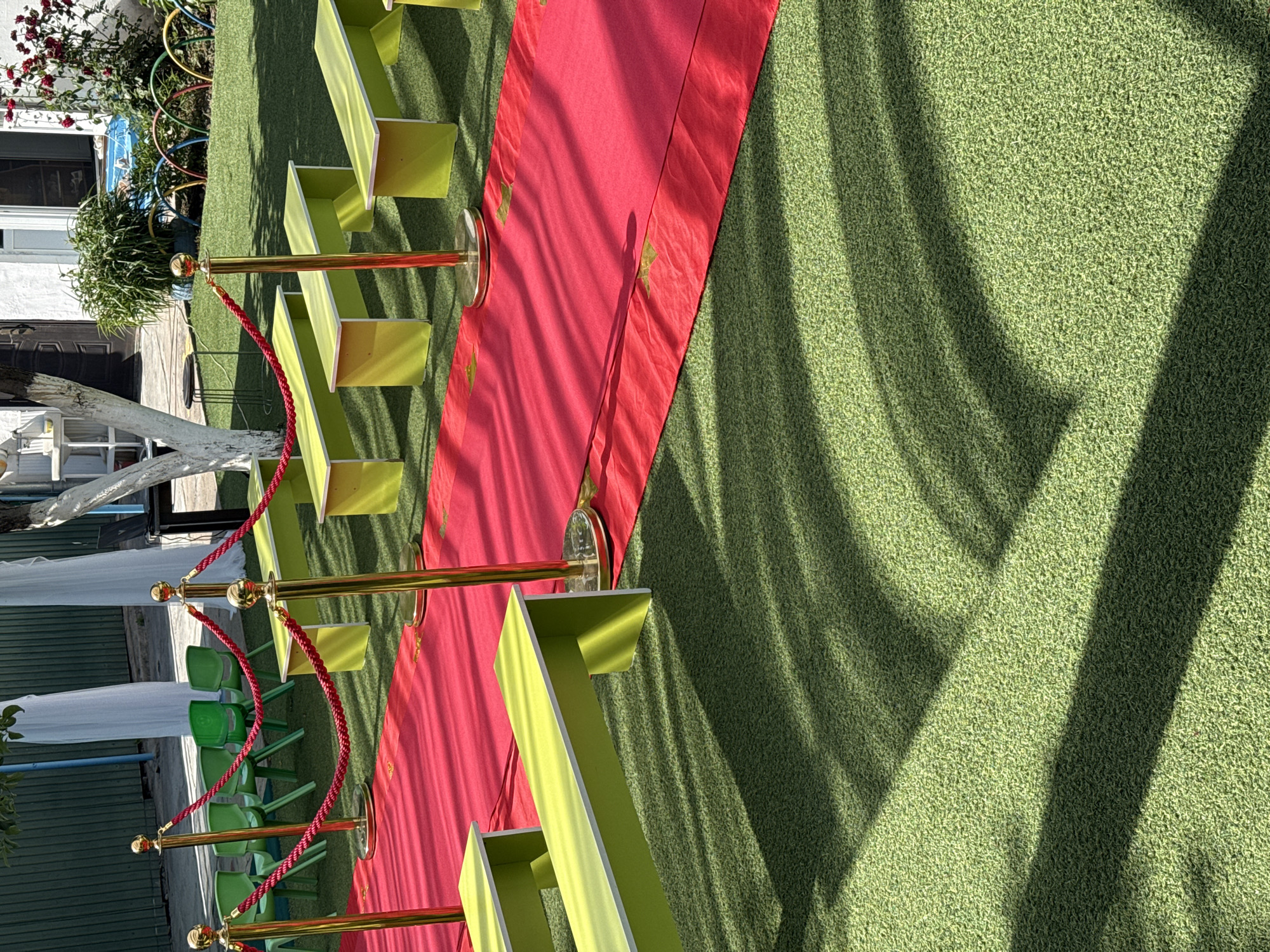 The image presents an outdoor scene featuring a red carpet runner flanked by yellow benches, positioned on a green grassy surface. The scene is bordered by gold stanchions connected with red rope. The location appears to be a garden or event space with dappled sunlight creating shadows across the grass. A building with windows and plants is visible in the background.
