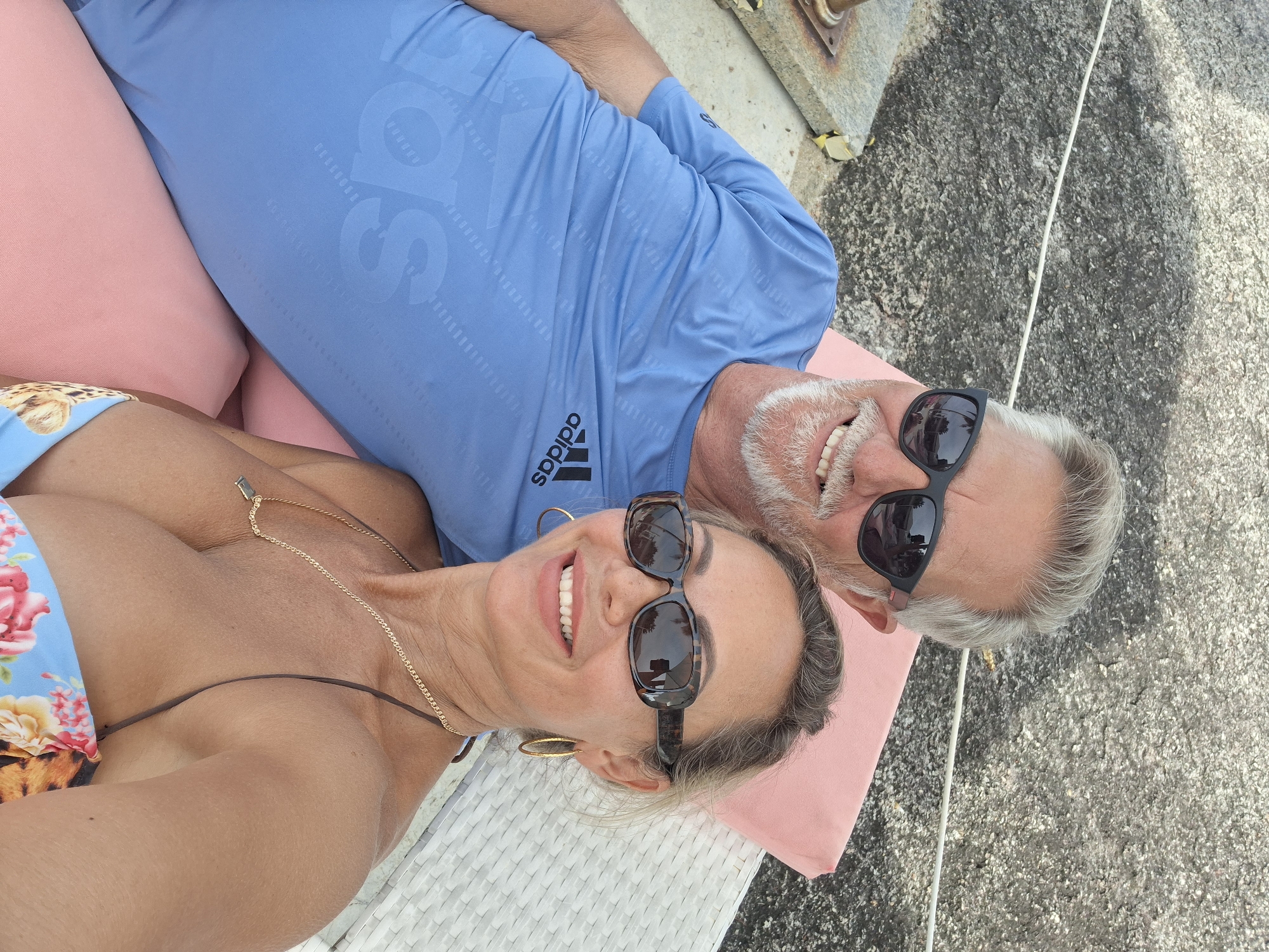 In this close-up, high-angle, outdoor selfie taken in Baan Hua Thanon, Thailand, a middle-aged couple lies on pink cushions on a textured grey surface, possibly a patio or deck. The man, on the right, wears a light blue long-sleeved t-shirt with a repeating Adidas logo pattern. He has grey hair and a beard, and is wearing dark sunglasses. His mouth is open in a wide smile, revealing white teeth. The woman, on the left, wears a floral-patterned bikini top and a gold necklace. She has on dark, tortoiseshell sunglasses and large hoop earrings. She is also smiling broadly, her teeth showing. The lighting suggests it is daytime, with the sun casting shadows on the ground. The overall mood is one of happiness and relaxation.