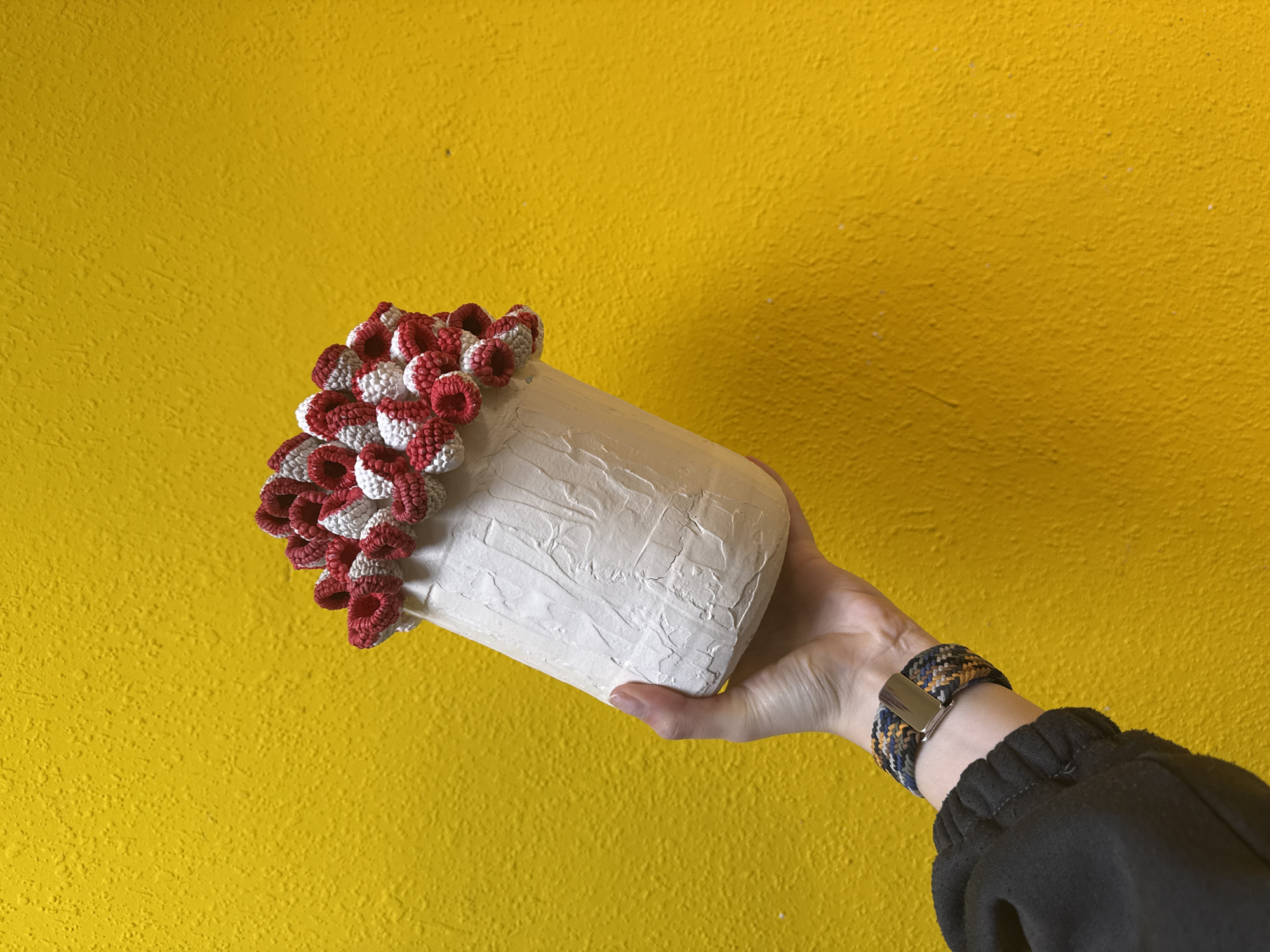 A hand holds a white cylindrical object with red and white crocheted embellishments against a textured yellow background. The hand wears a multi-colored woven bracelet with a silver clasp and is partially covered by a black sleeve. The location appears to be indoors, with the focus on the hand and object rather than the setting.