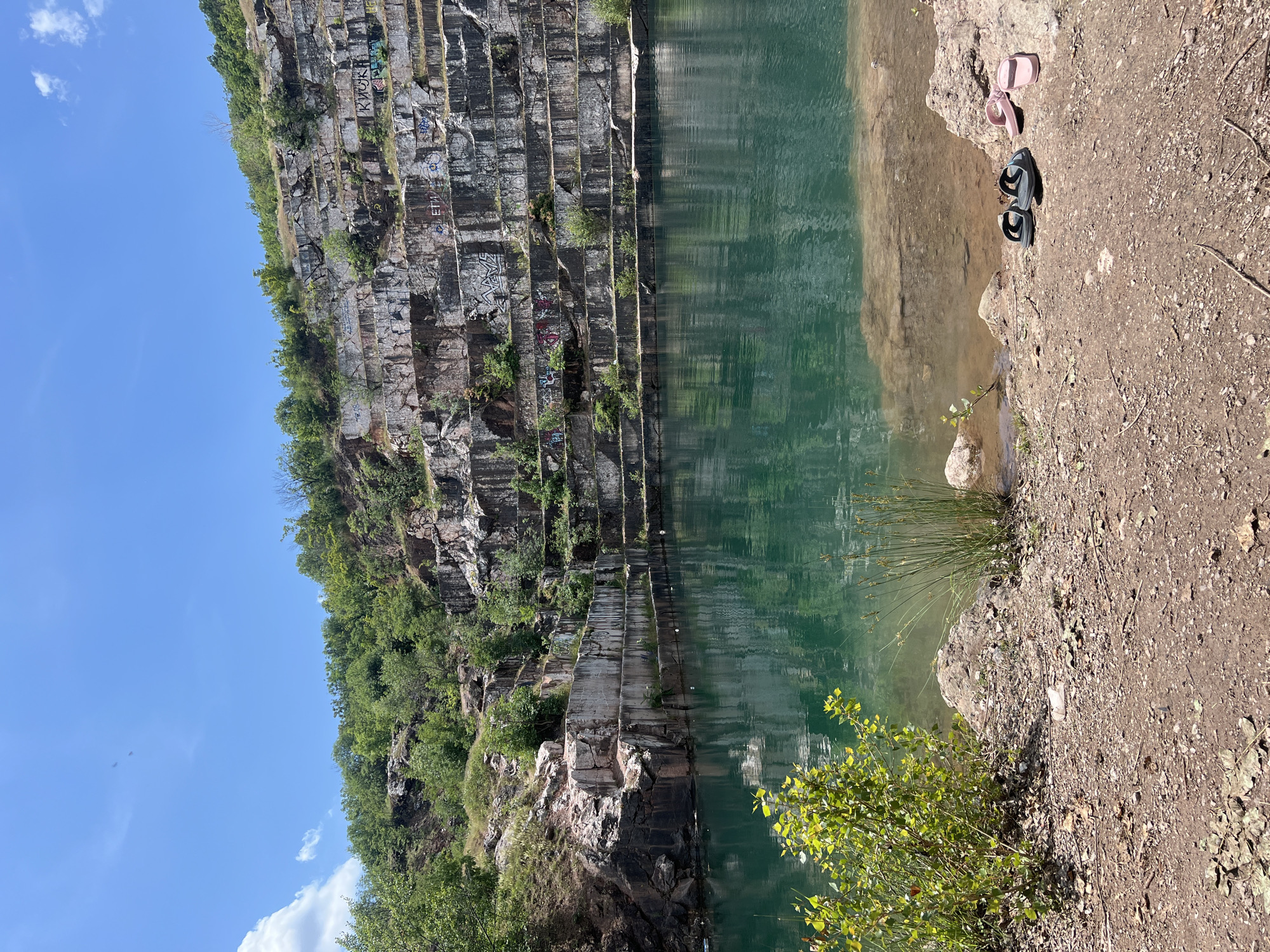 The image shows a quarry lake in Pravoslav, Bulgaria. The water is a deep turquoise color and reflects the surrounding rocky cliffs. The cliffs are layered and show signs of quarrying, with visible strata and some graffiti on the rock faces. Green vegetation grows on the upper parts of the cliffs and around the edge of the lake. In the foreground, on the rocky shore, there are two pairs of flip-flops: one black pair and one pink pair. The ground is covered in dirt, gravel, and fallen leaves. The sky is blue with a few scattered white clouds.
