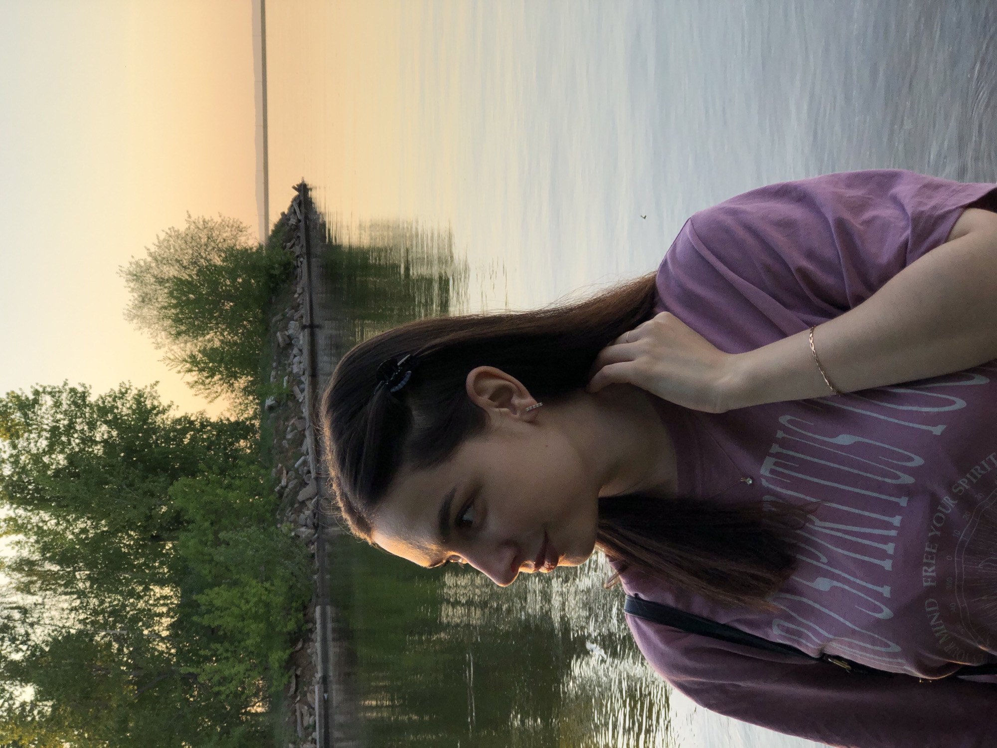 A young woman with long, dark hair stands near a body of water, likely the Kama River. The location is near Chaikovsky, Russia, based on visual cues. The woman wears a mauve t-shirt with white lettering. She has a bracelet on her left wrist and a hair clip in her hair. Her right hand is near her neck. Trees are visible in the background along the riverbank. The sky shows hues of orange and yellow, suggesting either a sunrise or sunset. The water reflects the colors of the sky. A strip of land with rocks is visible on the horizon.