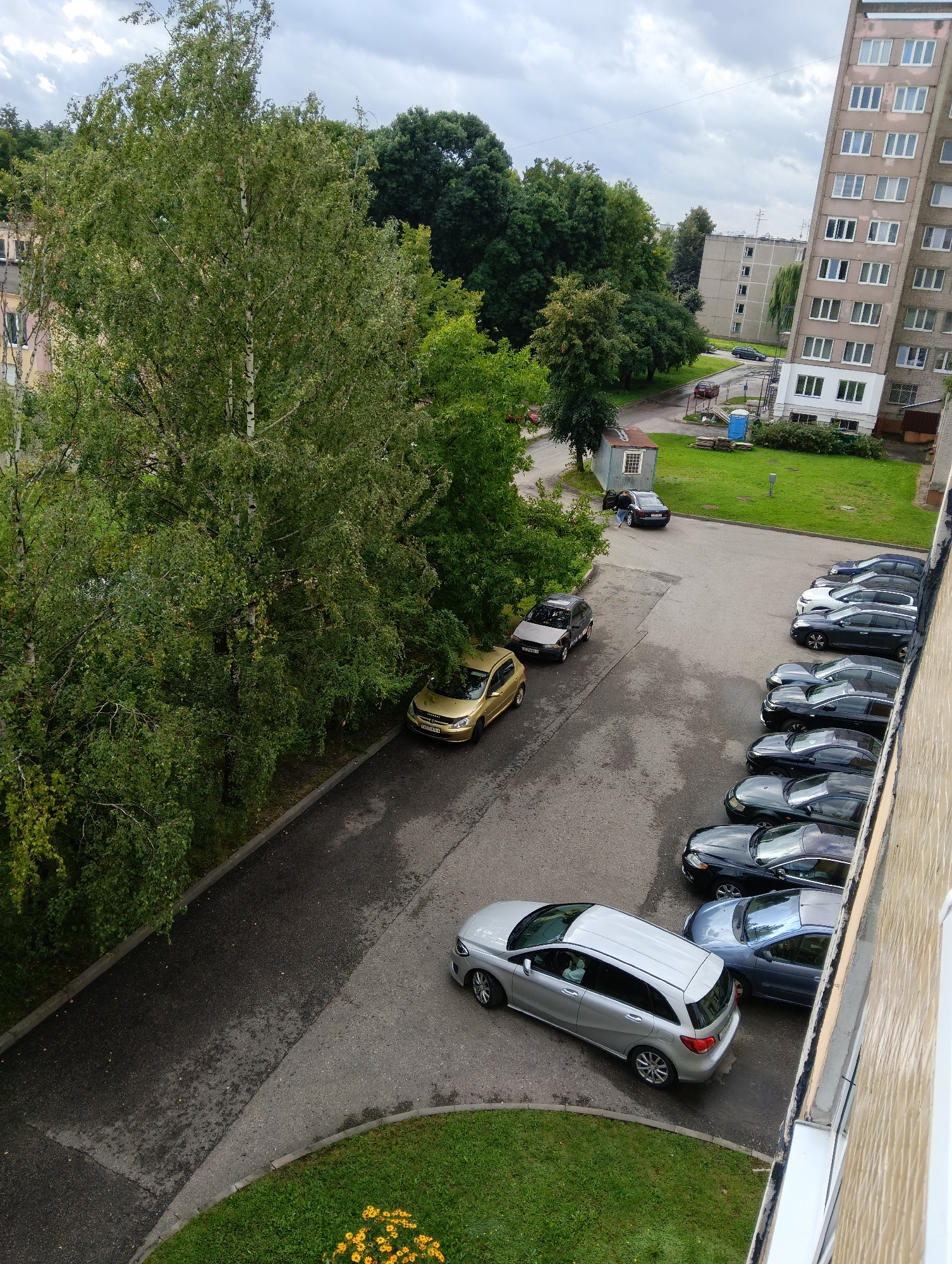 The image depicts an exterior scene in the city of Hrodna, Belarus, focusing on a parking lot adjacent to a tall residential building. The time of day appears to be daytime, with overcast skies.
In the foreground, a silver hatchback car is parked on the asphalt. To its left, a narrow strip of green grass with yellow flowers is visible. A portion of the textured, light-colored exterior wall of a building occupies the right edge of the frame.
Beyond the silver car, the parking lot continues, lined with several other vehicles. These include a dark blue sedan, a white sedan, a silver SUV, a black sedan, a dark gray sedan, a black sedan, and another dark gray sedan. Some of these cars are parked in a row, while others are arranged more individually.
Further into the scene, a gold sedan is parked on the side of a road, partially obscured by trees. To the left of the gold sedan, a dark-colored hatchback is parked, with its driver's side door open and a person visible inside.
Behind the parked cars, a small, light blue portable toilet is situated on a patch of green grass. Adjacent to the toilet is a small, weathered blue utility shed with a single window. A larger, grey residential building with numerous white-framed windows is visible in the background, along with other trees and buildings, suggesting a dense urban or suburban environment. The overall setting is a typical residential area.