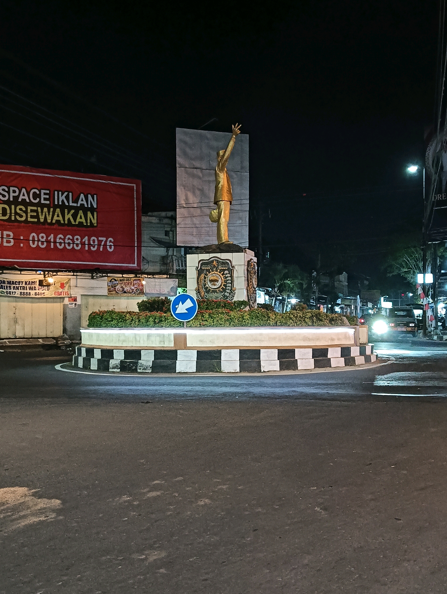 A golden statue of a figure with an outstretched arm is the central element of a roundabout in Purworejo, Indonesia. The statue stands on a decorated circular base with a tiered white and black checkered pattern. Lush green plants surround the base, and a blue circular sign with a white arrow indicating a left turn is placed on the island. In the background, a large red banner with white and yellow text reading "SPACE IKLAN DISEWAKAN" and a phone number is visible, along with other signs and buildings. Streetlights illuminate the scene, and a vehicle with its headlights on is visible in the distance, navigating the roundabout. The road surface is asphalt, and the overall impression is of a night-time urban environment.