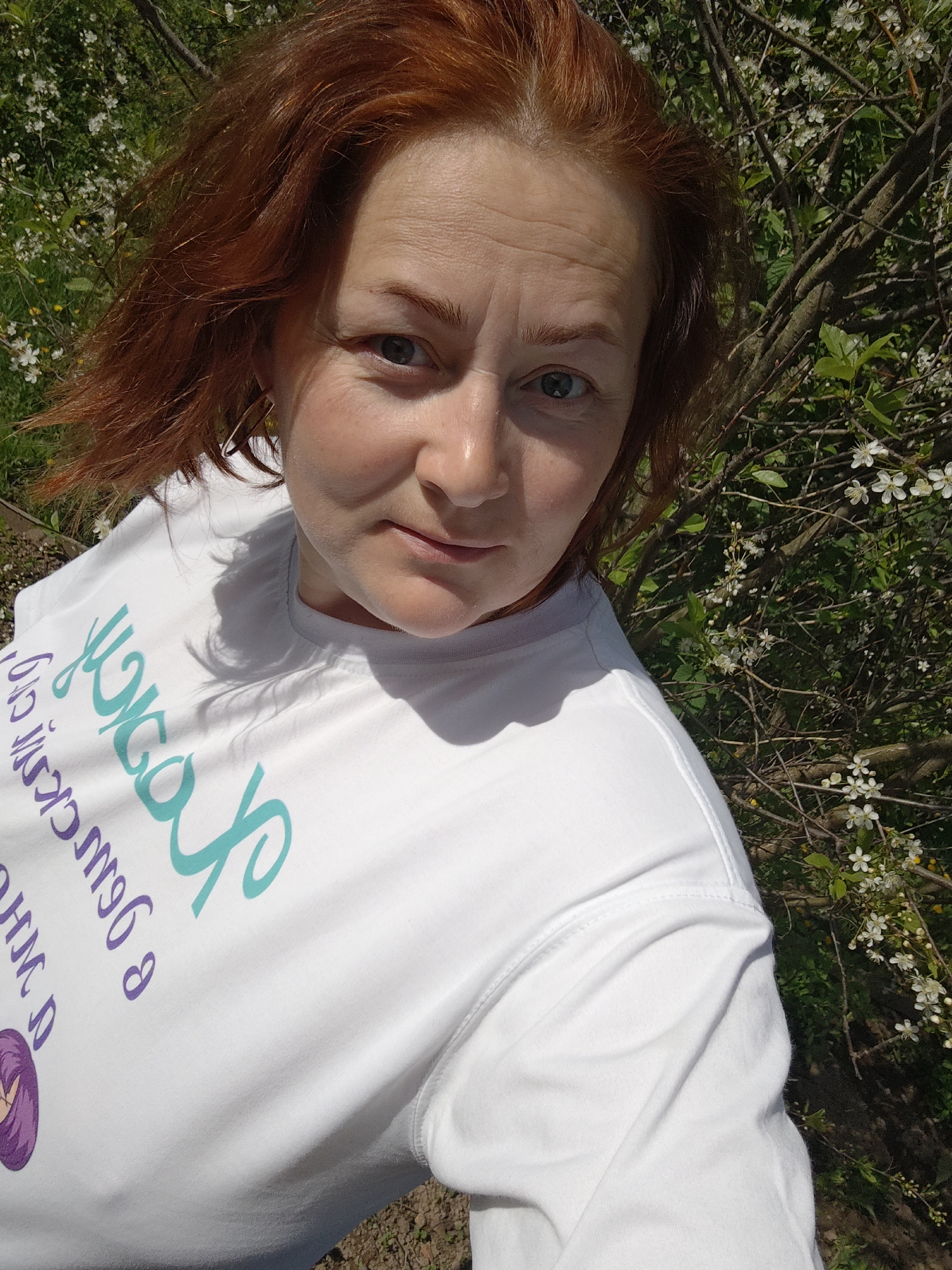 A woman with red hair poses for a selfie outdoors. She wears a white t-shirt with teal and purple writing. Behind her is a flowering bush with white blossoms and green leaves. The ground is visible near the bottom of the frame.
