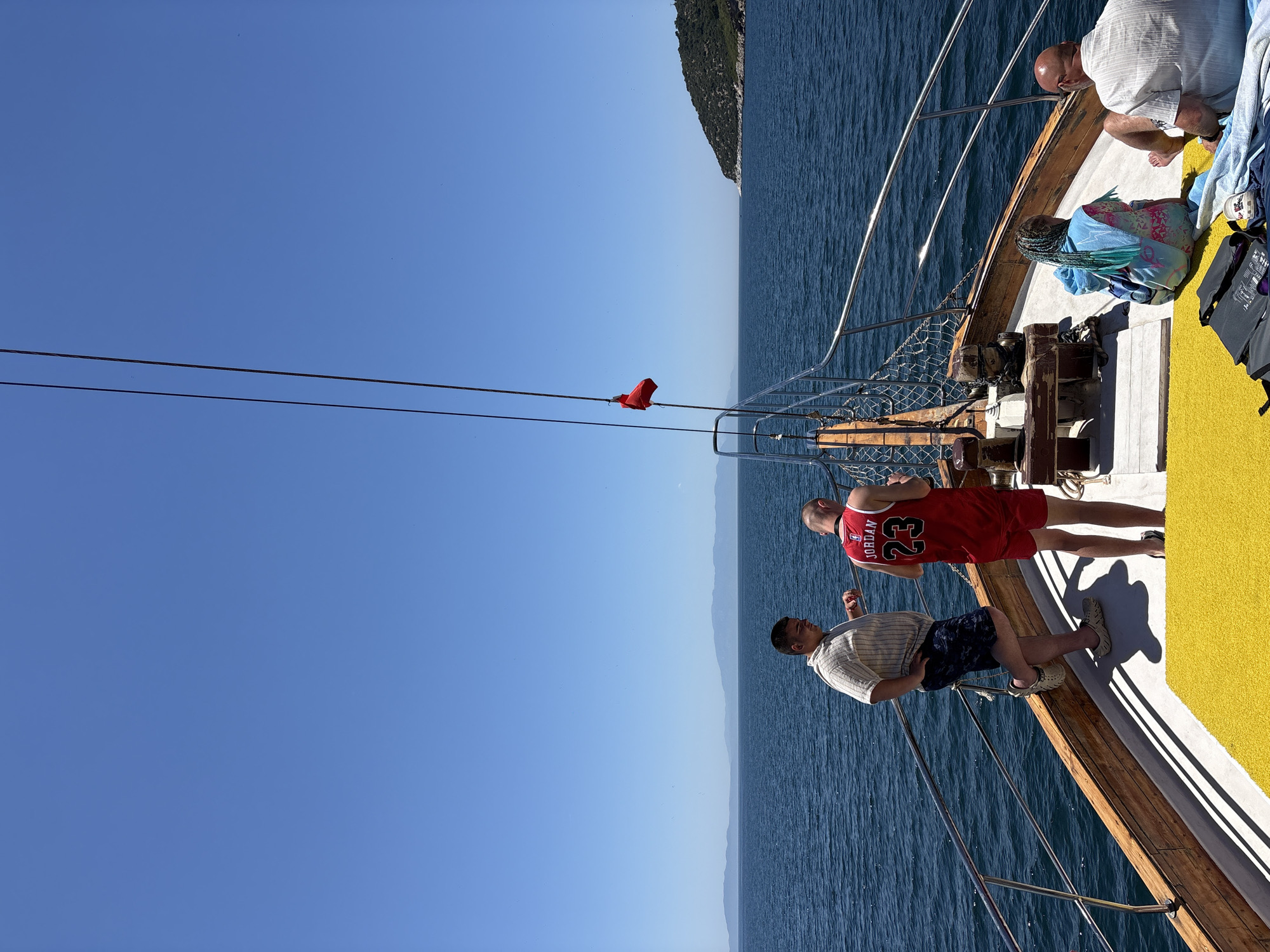 The image captures a sunny day on a boat trip near Menderes, Turkey, with clear blue skies and calm, deep blue water. The photograph is oriented vertically, showing the horizon running up the right side of the frame.
On the wooden deck of the boat, several individuals are present. In the foreground, two men stand. One man, positioned slightly to the right, wears a red basketball jersey with "JORDAN" and the number "23" printed in white. Next to him, another man wears a light-colored striped short-sleeved shirt, patterned shorts, and light-colored clogs. Further back on the deck, an older bald man wearing a white shirt is seated, looking towards the water. Nearby, another person with braided hair is partially visible, wrapped in a blue and pink patterned blanket.
The boat deck features a yellow matting covering part of the surface, contrasting with the natural wood and white sections. Metal railings run along the side of the boat, separating the deck from the sea. Ropes or cables stretch across the frame, with a small red flag or piece of fabric attached to one in the mid-ground.
In the distance, the shoreline of hills or mountains is visible under a bright, cloudless sky, suggesting a coastal area. The strong sunlight casts shadows from the boat's structures and the people. On the deck, blue and white towels are casually placed, and a black bag with "NORTH FACE" visible on it, along with a white cup, are also present. The scene conveys a relaxed, leisurely atmosphere on the water during daytime.