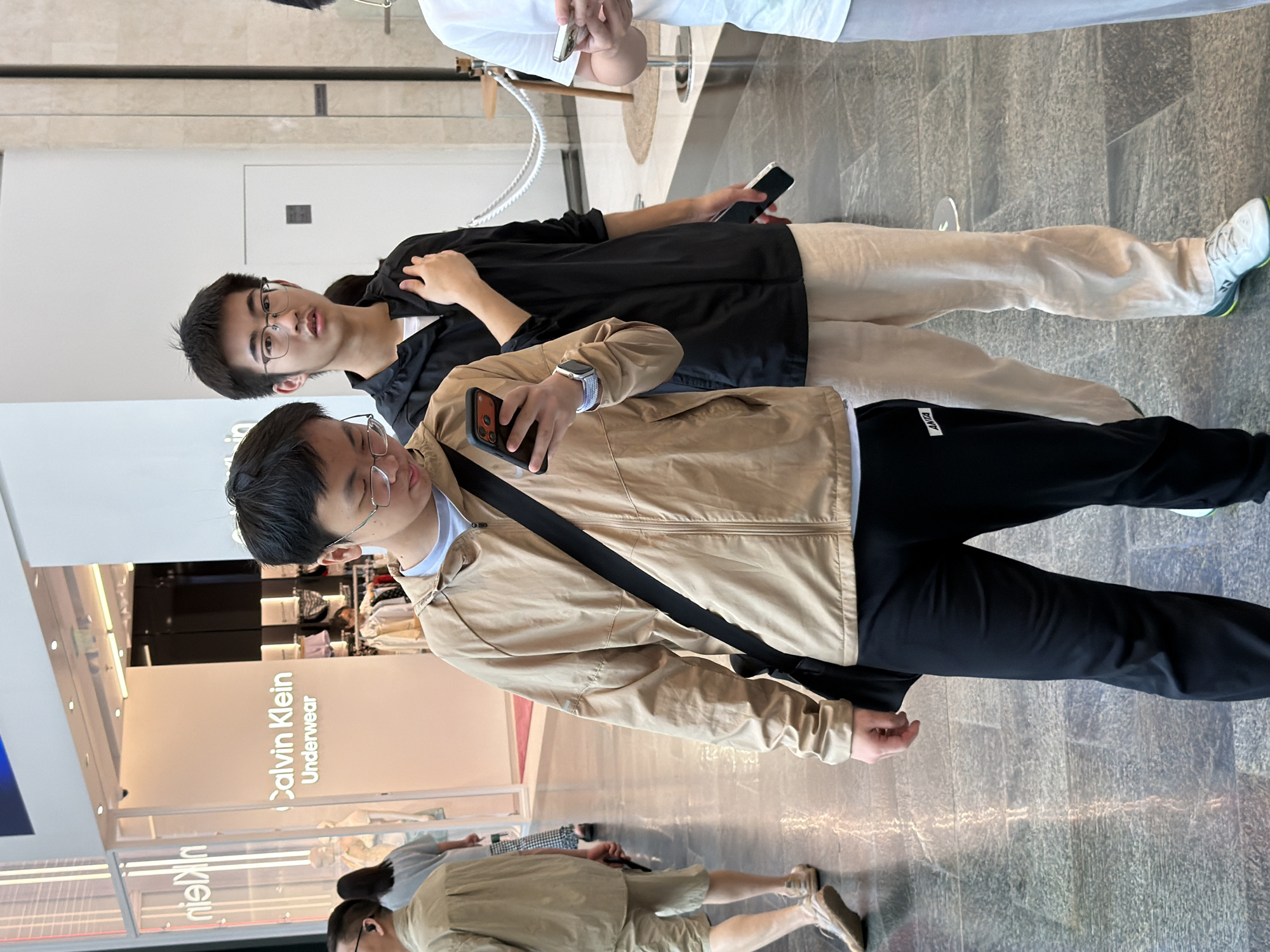 The image depicts an indoor scene, likely a shopping mall, with a Calvin Klein Underwear store visible in the background. Three young men are the central subjects. The man in the foreground, on the left, is wearing a tan jacket, black pants, and holding a smartphone. To his right stands another man in a black jacket and tan pants, looking towards the camera and holding a smartphone. Further to the right, a third man in a white t-shirt and grey pants is partially visible, also holding a smartphone. The flooring is made of polished stone tiles. The overall setting suggests a commercial environment within Yuzhong District, China.