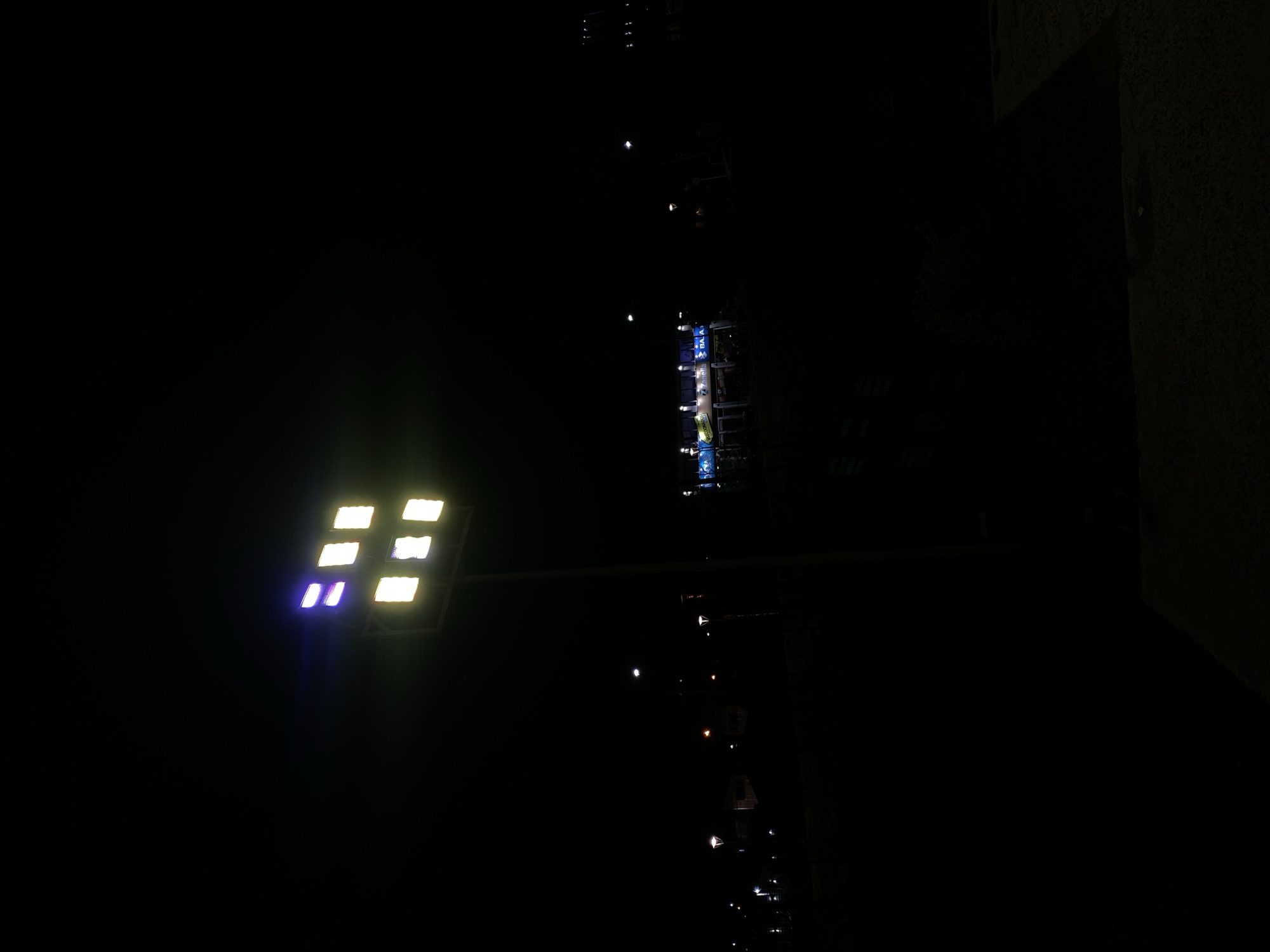 The image depicts a night scene in Ushuaia, Argentina. A bright rectangular LED street light, with a matrix of individual lights, illuminates the foreground from the left. The upper right is notably dark, potentially a building corner. Further away, along the line of street lamps, a building with blue illuminated signage is barely visible in the distance. The general environment is dark, with minimal ambient light.