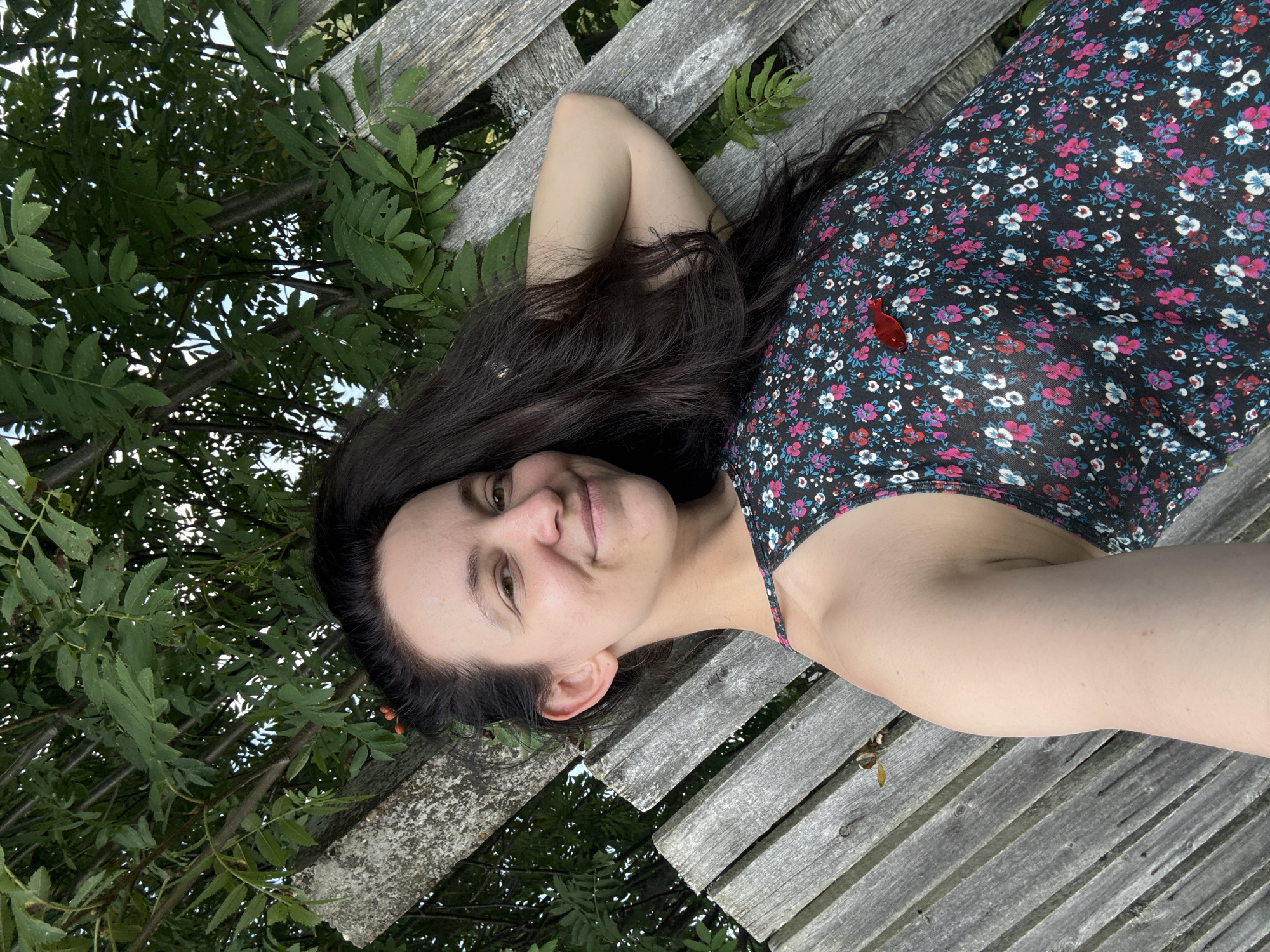A young woman with long, dark hair smiles at the camera, leaning back against a rustic wooden fence. She is wearing a dark, floral-patterned dress with thin straps. Her right arm is bent, with her hand resting on the back of her head. Green foliage from a tree is visible behind and above her, with some sunlight filtering through the leaves. The fence is made of weathered, grey wooden planks arranged diagonally. The scene appears to be outdoors, in a natural setting. There are no visible indications of Zasechnyi, Russia.
