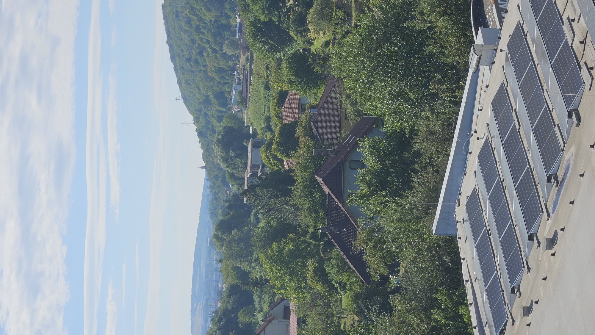The image captures an aerial view of a suburban or semi-rural area, likely within or on the outskirts of Prešov, Slovakia. In the foreground, a flat, light-colored roof is covered with multiple rows of dark, rectangular solar panels, arranged in a grid pattern. These panels appear to be angled slightly upwards, suggesting a setup for optimal sunlight absorption. The roof also has several circular vents and some black conical fixtures. Beyond the solar panel-covered roof, a dense expanse of lush green trees dominates the middle ground, interspersed with several residential buildings. Some of these houses have visible tiled roofs in shades of brown and red, and one building in the foreground, partially obscured by trees, displays a light blue facade. Further in the distance, a hilly, tree-covered landscape rises, with hints of more scattered buildings and infrastructure, including what appear to be power lines and a pylon on the highest hill. The sky above is a pale blue, with thin, horizontal white clouds streaking across it. The overall impression is one of a sunny day in a verdant area, where residential development coexists with significant natural greenery.