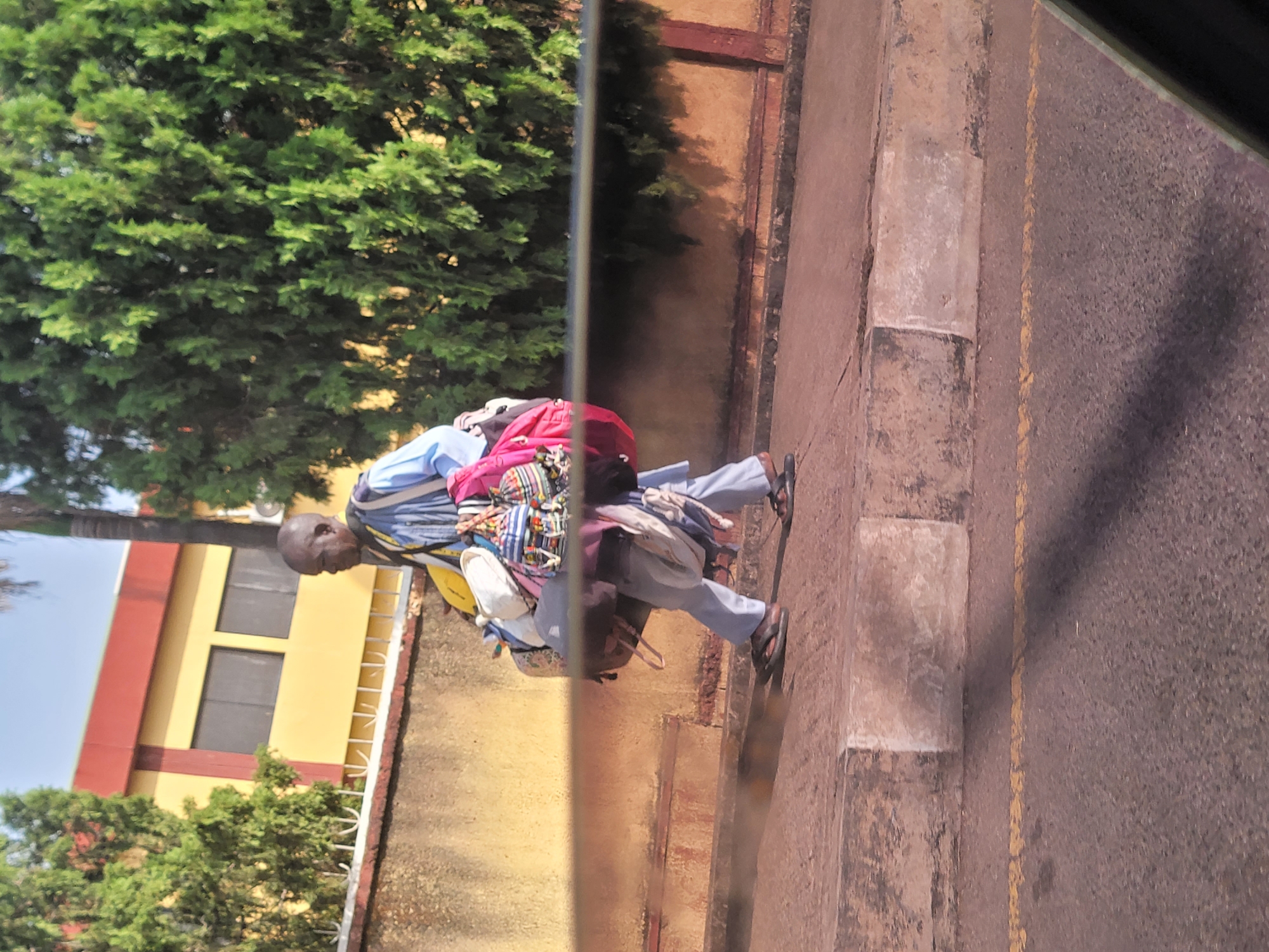 A man is walking on a sidewalk next to a road. He is heavily burdened with luggage and bags of various sizes and colors. The man is wearing a light blue shirt and dark sandals. The sidewalk is made of concrete blocks and is next to a paved road marked with a yellow line. Behind the sidewalk and road, there is a wall with a building visible above it. The building is painted in yellow and red. Green trees are visible in the background. The location is Jos, Nigeria.
