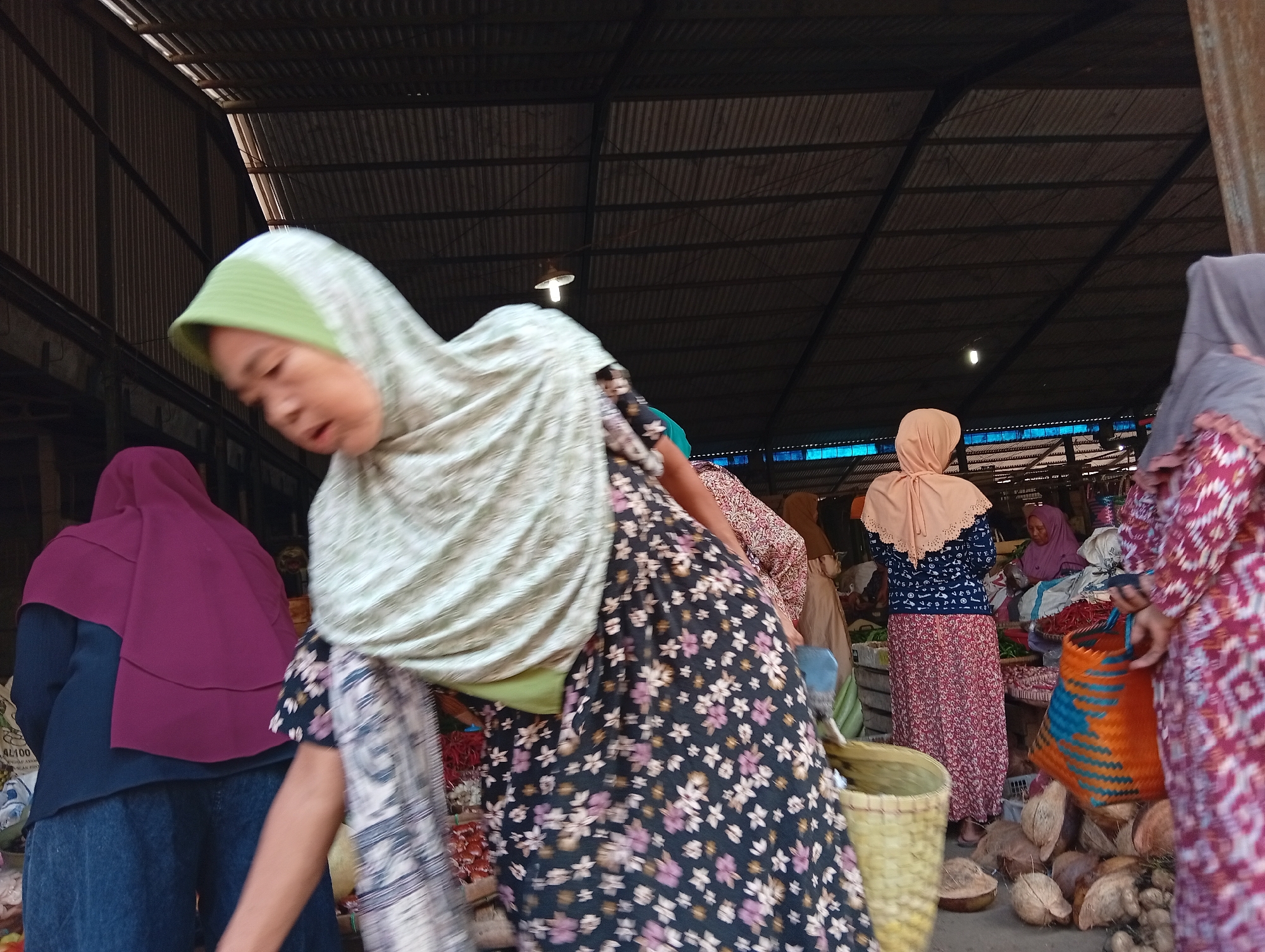 The image depicts an outdoor market scene in Plempukankembaran, Indonesia. In the foreground, a woman wearing a green and white patterned hijab and a black floral-patterned dress is partially visible, leaning forward. To her left, another woman is seen from the back, wearing a dark blue top and a purple hijab, with denim pants. In the background, several other individuals are present. One woman in a peach-colored hijab stands near a stall laden with items, possibly produce. Another woman, in the far right, is wearing a maroon patterned dress and holding an orange woven bag. The market appears to be covered by a corrugated metal roof supported by visible metal beams, suggesting a semi-permanent structure. The ground is covered with a mix of debris and what appear to be coconuts. The overall atmosphere suggests a bustling local marketplace.