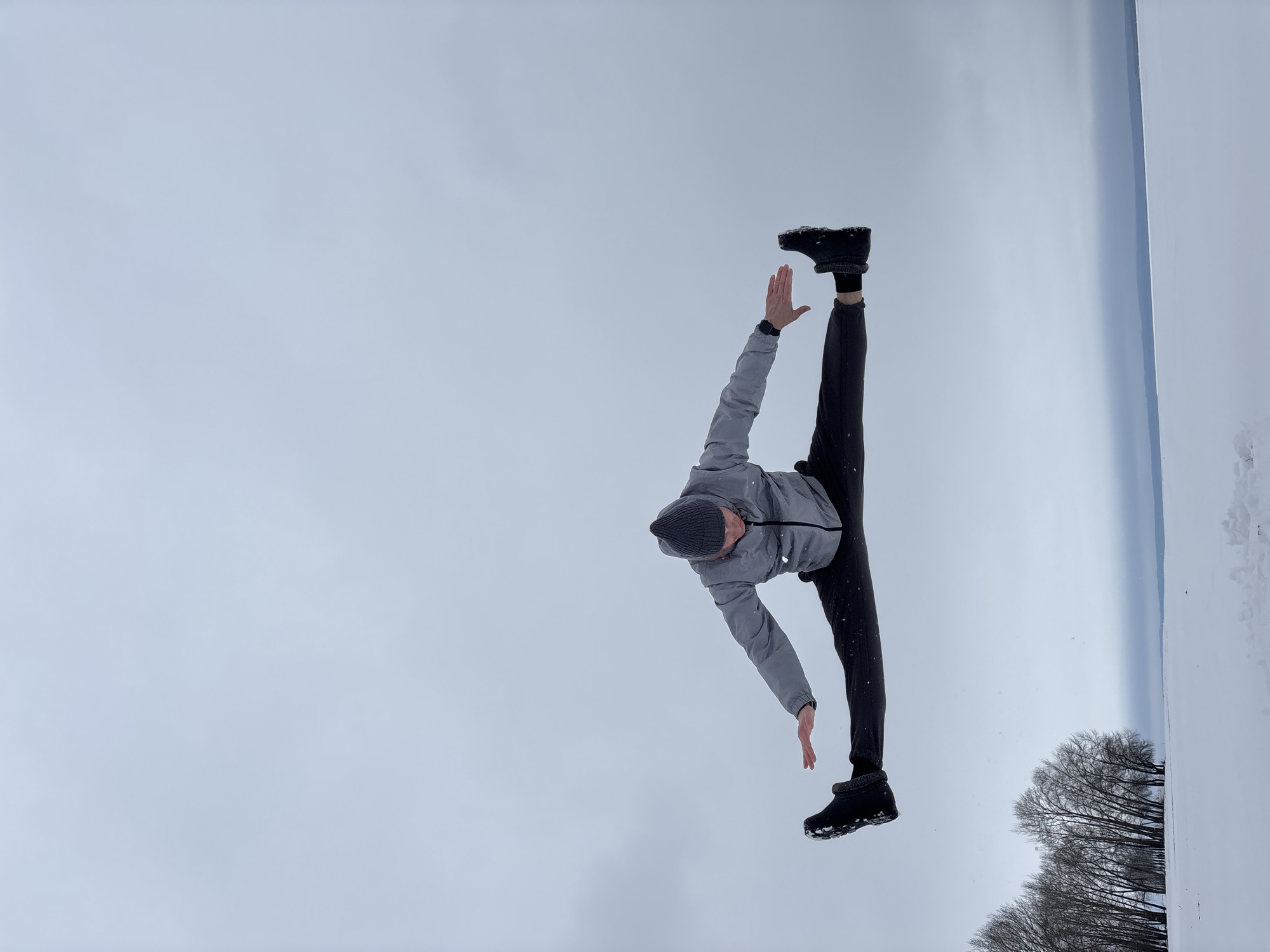 A man in a gray jacket, black pants, and a gray beanie is performing a split in a snowy outdoor environment. He is holding his arms out to the sides, one hand raised, and the other pointing downwards. The background consists of a snow-covered field and some bare trees. It is an overcast day. The location is Noviy, Russia.