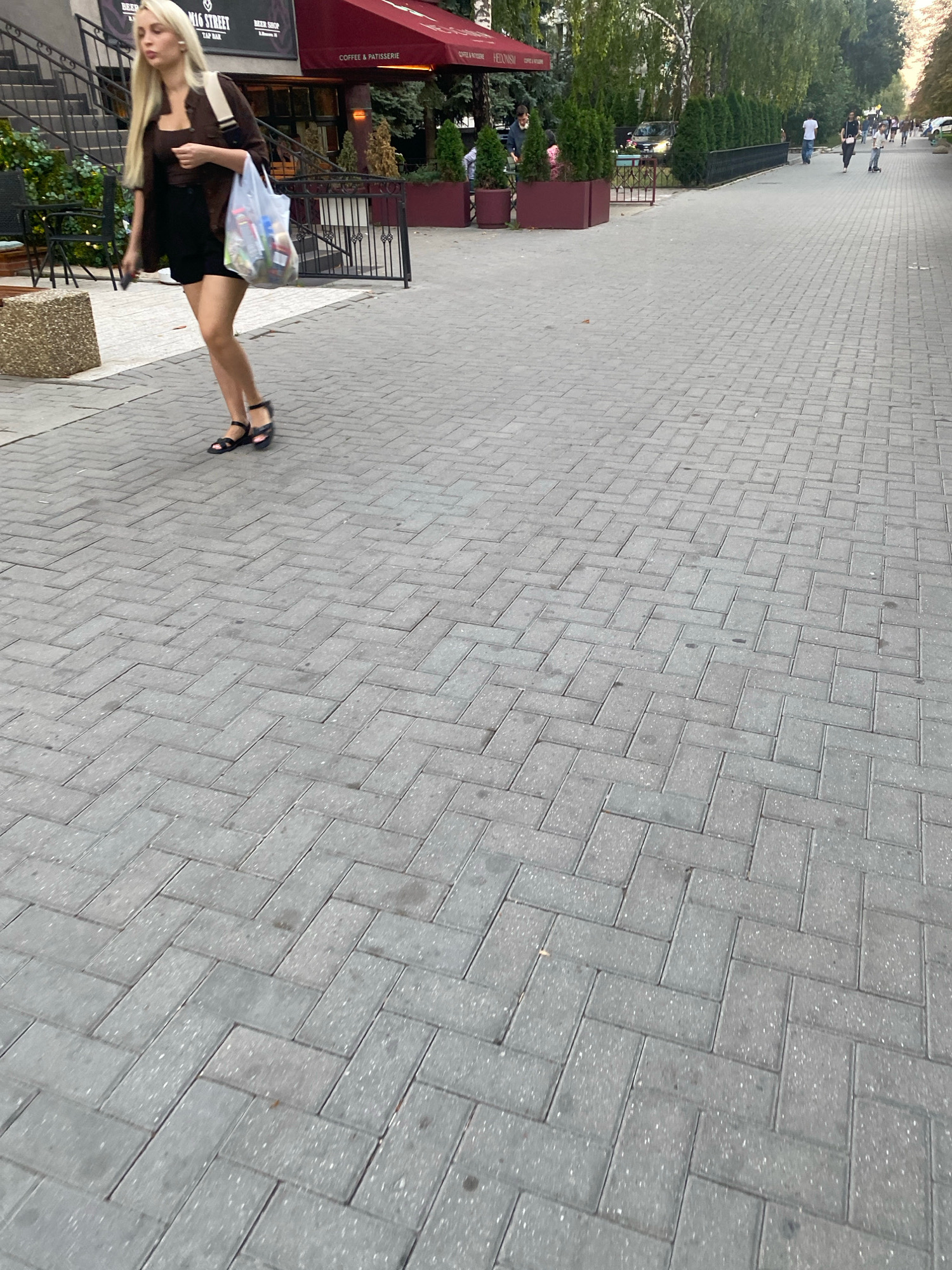 The image shows a young woman with blonde hair walking on a pedestrian sidewalk made of gray rectangular pavers laid in a herringbone pattern. She is wearing a dark brown short-sleeved shirt, black shorts, and black sandals. She is carrying a transparent plastic bag filled with various items in her right hand and a dark object, possibly a phone, in her left hand. Behind her, a cafe with a red awning and outdoor seating is visible, with people dining and walking. In the background, trees and other pedestrians are present on a wider sidewalk area, with cars parked further down the street. The setting appears to be an urban environment with a mix of commercial and residential buildings. The specific location is Stefan cel Mare Boulevard in Chișinău, Moldova, identifiable by the cafe's sign which reads "COFFEE & PATISSERIE" and the general urban architecture visible.