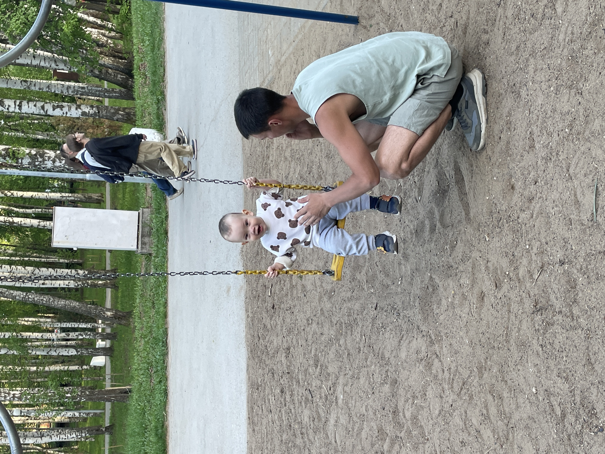 A man kneels and looks at a baby on a swing in a park. The park has many birch trees. In the background, two people stand on a sidewalk. This photograph was taken in the city of Perm, Russia.