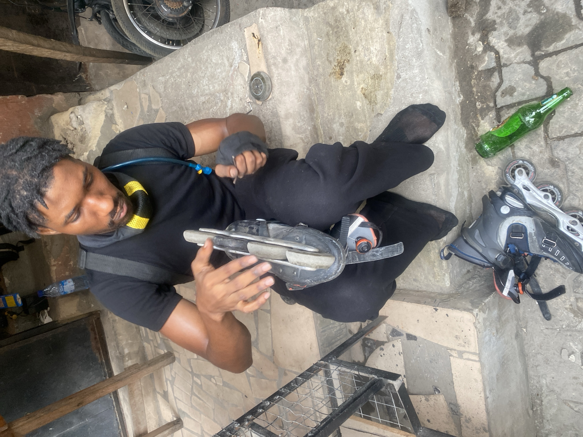 A man wearing black clothing and a hydration pack is sitting on concrete steps, holding an ice skate in his hands. A pair of inline skates, a green glass bottle, and a loose skate wheel are also on the steps. A bicycle wheel is visible in the background, along with metal framing. The image appears to be taken in a somewhat cluttered, outdoor space, possibly in Lagos, Nigeria.