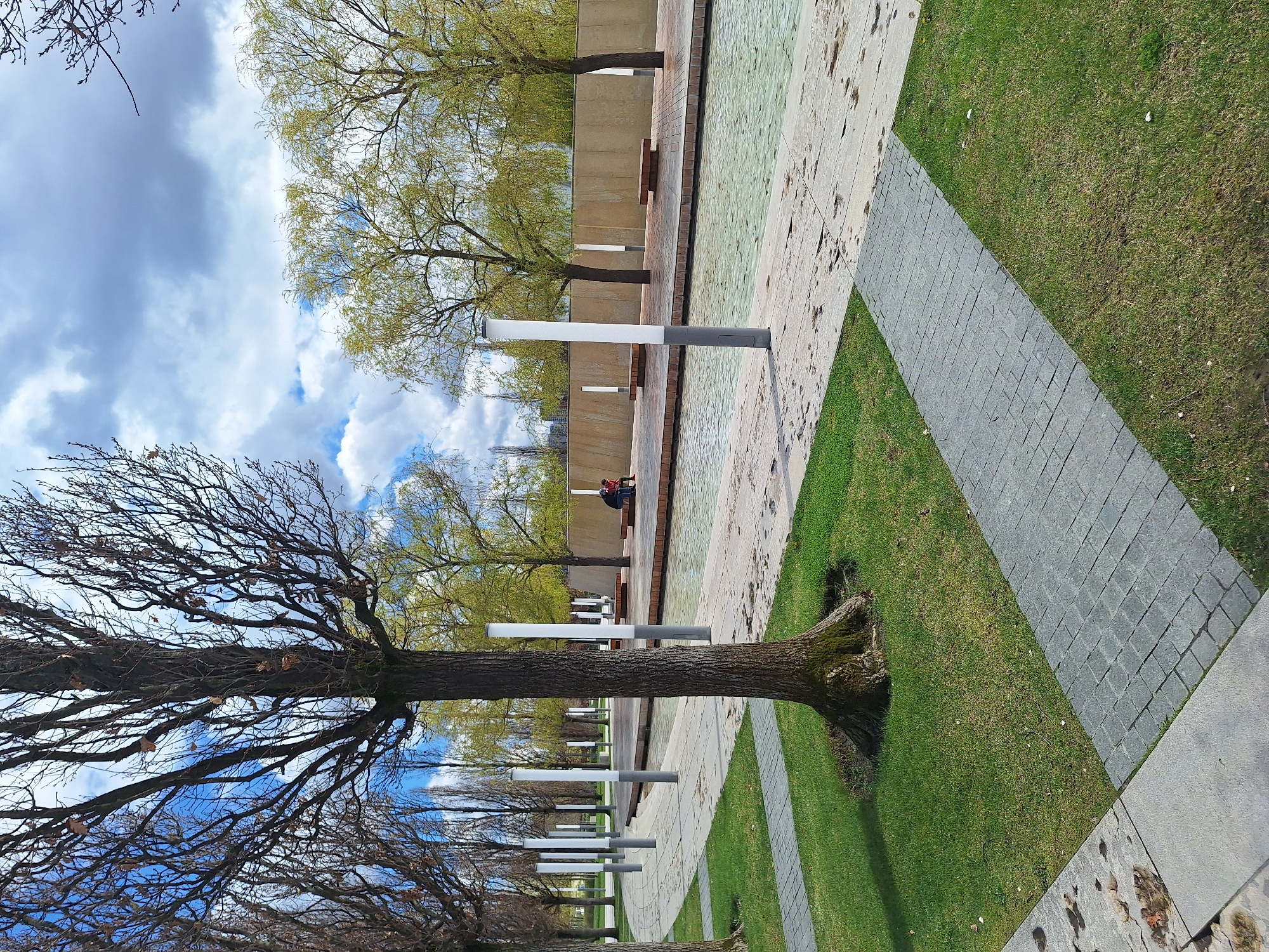 The image shows a scene in what appears to be a public park or memorial in an urban setting. A row of deciduous trees lines a walkway. Some trees have no leaves, while others are budding with new foliage. Adjacent to the walkway is a rectangular area of paving or concrete that appears to be a water feature with a layer of clear water.
Along the edge of this water feature are regularly spaced white and gray lamp posts. In the background, two people are sitting on a ledge by a concrete wall. The sky overhead is partially cloudy. The trees are planted in patches of grass, bordered by gray brick pavers.