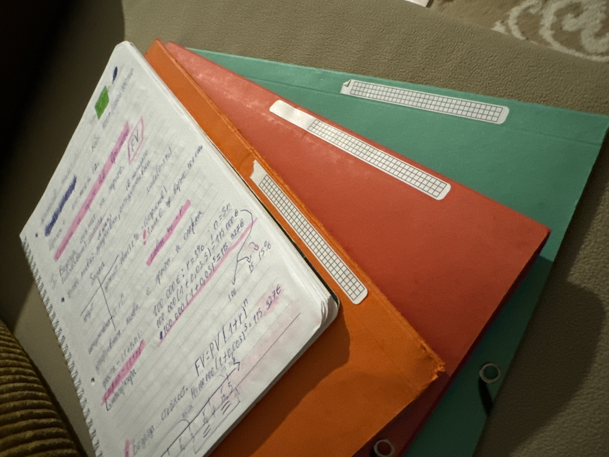 The image shows a notebook resting on a stack of colored folders. The notebook is open, revealing handwritten notes with mathematical formulas and financial concepts, some highlighted in pink. The folders are orange, red, and green, with white label areas on each. The objects are resting on a textured surface, possibly a couch or a rug.