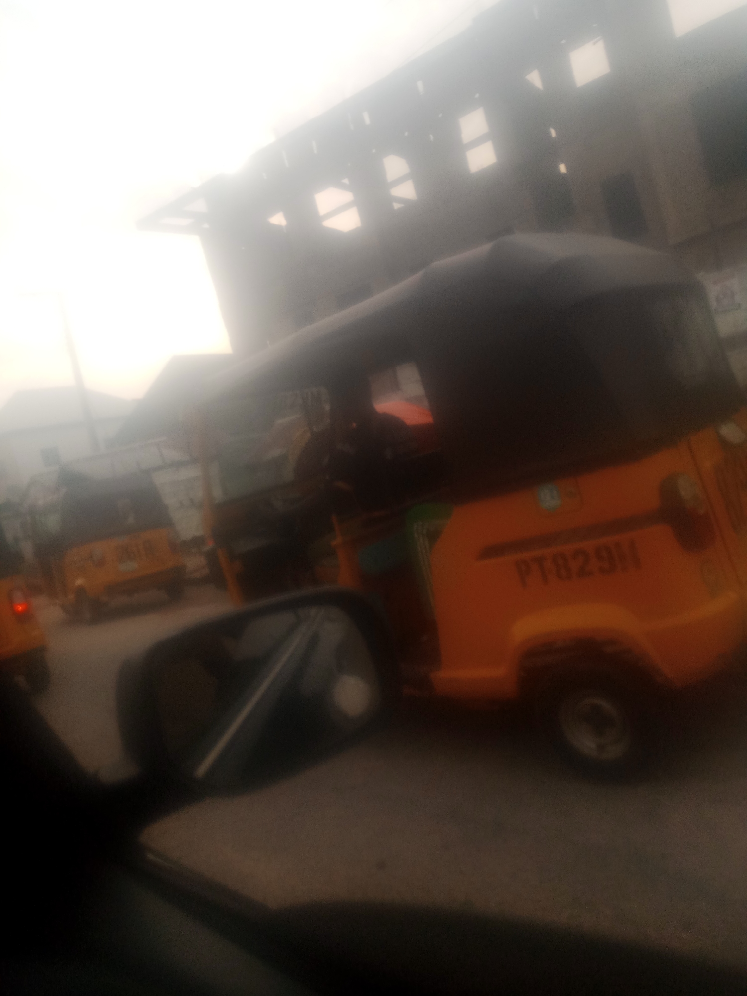 The image depicts a street scene in Jalingo, Nigeria, characterized by a hazy atmosphere. The foreground shows the interior of a vehicle, likely a car, with a side mirror visible. The primary subjects are several motorized rickshaws, commonly known as "Keke Napep" in Nigeria. One prominent Keke Napep, orange in color with a black canopy, displays the license plate "PT82911". Another yellow Keke Napep with the partially visible license plate "261P" is positioned to the left. The background is dominated by a blurred building with a distinctive architectural style, featuring a series of rectangular window-like openings. The overall impression is of a typical urban environment in Jalingo, likely during daylight hours, with moderate traffic and limited visibility due to atmospheric conditions.