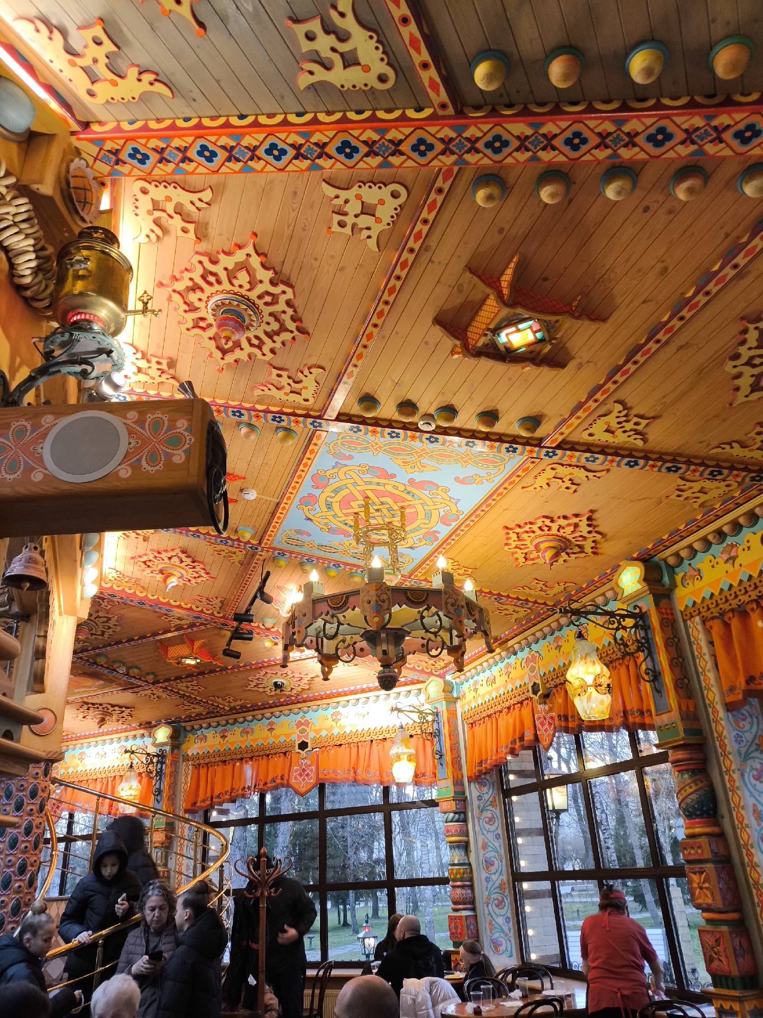 The image captures the vibrant and elaborately decorated interior of a restaurant or cafe, possibly with a traditional Russian folk art theme. The most striking feature is the wooden ceiling, extensively painted with colorful patterns in red, yellow, blue, and orange, featuring stylized floral motifs, geometric designs, and carved wooden ornaments resembling birds or abstract shapes. Multiple light fixtures are visible, including a large central chandelier with candle-like bulbs, smaller ornate hanging lanterns with colored glass, and decorative spherical lights on the ceiling.
On the left, a brass samovar and a wooden speaker box, also adorned with folk art designs, are suspended. Below, a spiraling staircase with a golden railing leads downwards, where several people, including a woman looking at her phone, are gathered. In the lower right, people are seated at tables, some conversing, while one person in a red shirt appears to be a staff member near a large window. The windows offer a view of an outdoor area with bare trees, suggesting autumn or winter.
The walls and pillars are also richly decorated with matching colorful patterns. Orange valances with decorative borders hang above the windows, further enhancing the festive and traditional atmosphere. The overall scene depicts a lively indoor gathering, likely during daytime or early evening, characterized by its intricate, handcrafted decor and warm lighting. No discernible text is visible in the image.