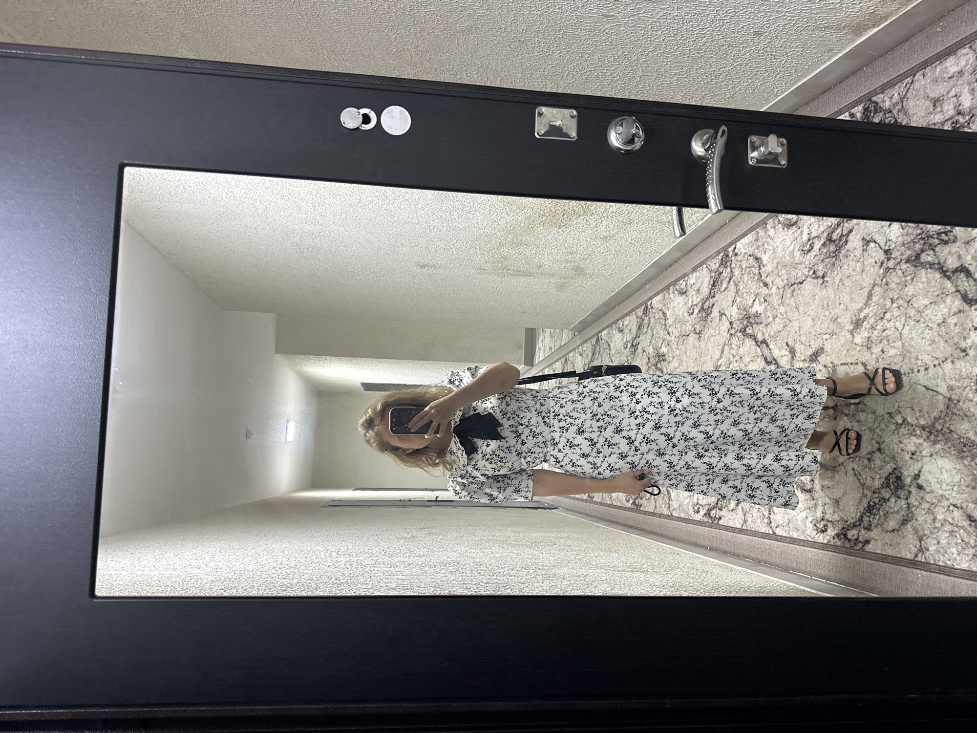 A woman in a white floral dress with short puff sleeves and a black bow tie at the neck stands in a hallway and takes a selfie in a full-length mirror. She is wearing black strappy high-heeled sandals. A black handbag is slung over her right shoulder. The hallway is narrow with textured light-colored walls. The floor is covered in a marble-patterned carpet. A black door with metallic hardware, including a handle and locks, is partially visible on the right side of the frame. The image appears to have been taken indoors, possibly in a residential building hallway, though no specific city or country information is ascertainable from the visual content alone.
