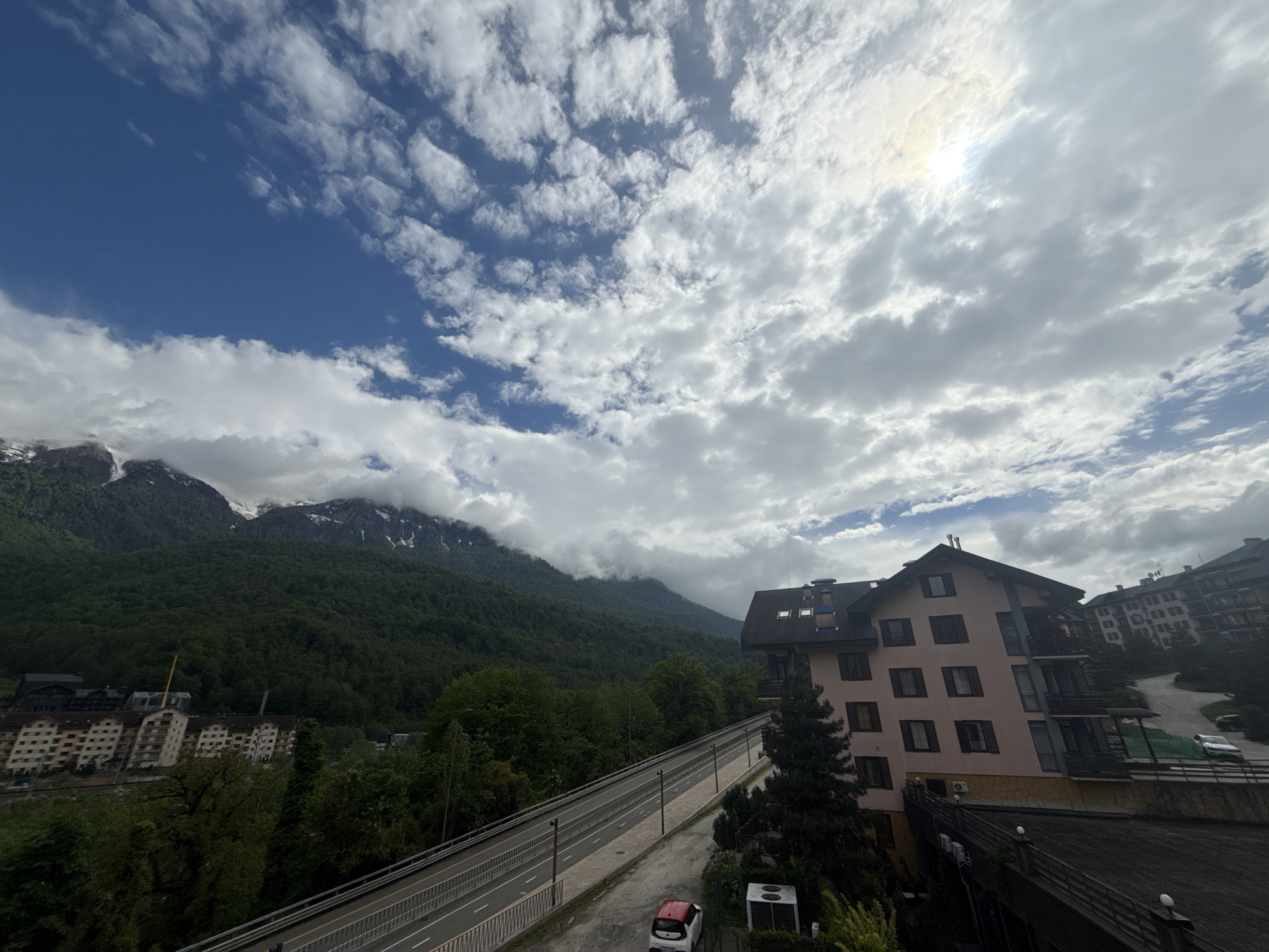 The image captures a view in Krasnaya Polyana, Russia. In the background, mountains are visible with some snow cover on their peaks. Lush green trees cover the lower slopes of the mountains. In the midground, there are several buildings, including a pink-colored building with dark shutters. A road runs parallel to this building. A white car with a red roof is parked near the road. The sky above is filled with clouds, with patches of blue sky visible. The sun is shining brightly, creating a glare. Other buildings are visible in the distance on the left side.