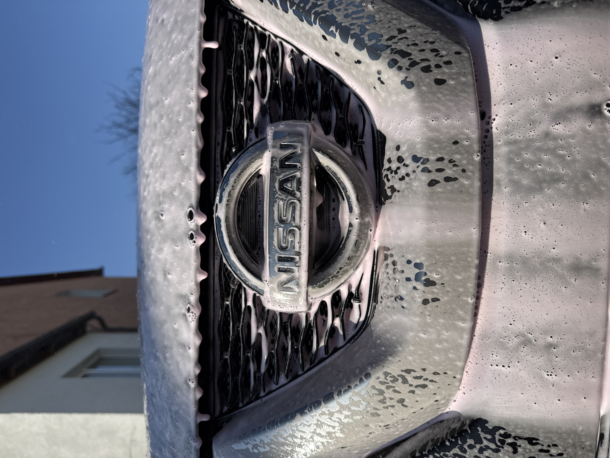 A close-up photograph shows the front grill and Nissan logo of a car covered in soapy foam. The grill is black with a silver Nissan logo in the center. Pinkish-white soapy foam covers the surface of the grill and surrounding areas. The background reveals a building with a white wall and a brown roof on the left, as well as a clear blue sky with a bare tree branch in the upper left corner. The location, Ждановіцкі сельскі Савет, Belarus, is irrelevant to the observable content of the image.