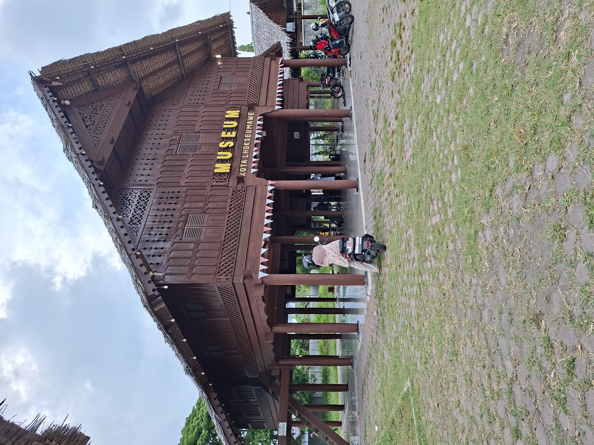 The image shows the Museum Kota Lhokseumawe in Lhokseumawe, Indonesia. It is a brown wooden building with a traditional design and a thatched roof. The word "MUSEUM" is displayed prominently in yellow letters above the entrance. There are several motorcycles parked in front of the museum. A person wearing a headscarf is standing near one of the motorcycles. The surrounding area appears to be a mix of paved surfaces and grassy areas. The sky is overcast.