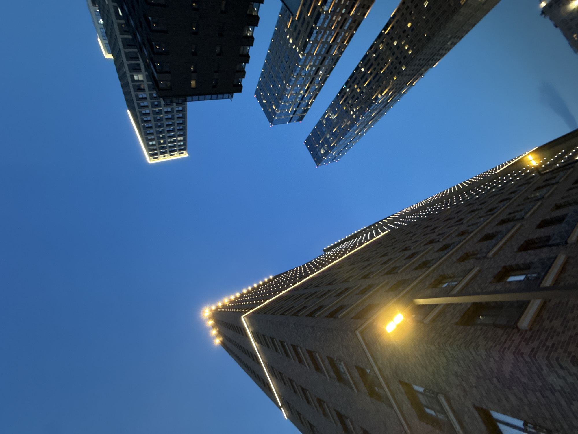 The image is an upward-angled photograph of several skyscrapers against a blue sky, taken in the city of Moscow, Russia. The skyscrapers are illuminated with lights along their edges and within some of the windows. The building on the bottom right appears to be made of brick and has a streetlight attached to its side. The other buildings in the image have a more modern, glass-and-steel design. There are no people visible in the image. The overall impression is one of urban density and modern architecture illuminated against the dusk sky.