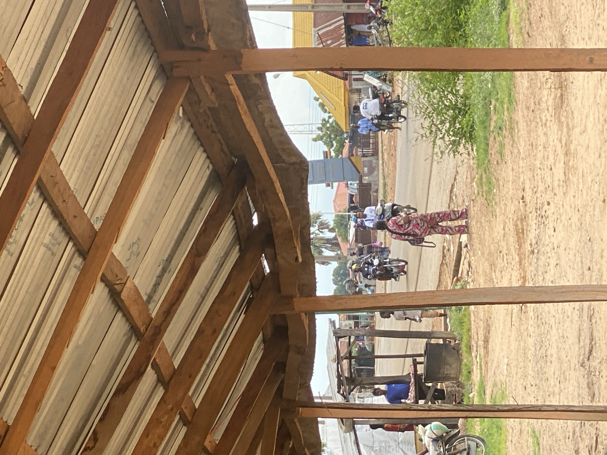In Bali, Nigeria, the image captures a street scene viewed from under a makeshift wooden shelter with a corrugated metal roof. In the foreground, two rough-hewn wooden posts support the shelter, with sandy ground beneath. A person in a pink and white patterned outfit stands in the middle ground, facing away from the camera. To the left, a man in a blue shirt stands behind a table displaying meat. A young boy in a white t-shirt stands near him, facing the street. Across the dusty road, several people are on motorcycles. One man in a white shirt with a black logo rides a motorcycle towards the viewer. Another man in a blue shirt is also on a motorcycle further down the road. Buildings with corrugated metal roofs, one with a bright yellow roof, line the street. Lush green vegetation is visible on the right side of the road, contrasting with the dusty ground. The overall impression is that of a bustling, everyday scene in a Nigerian town.