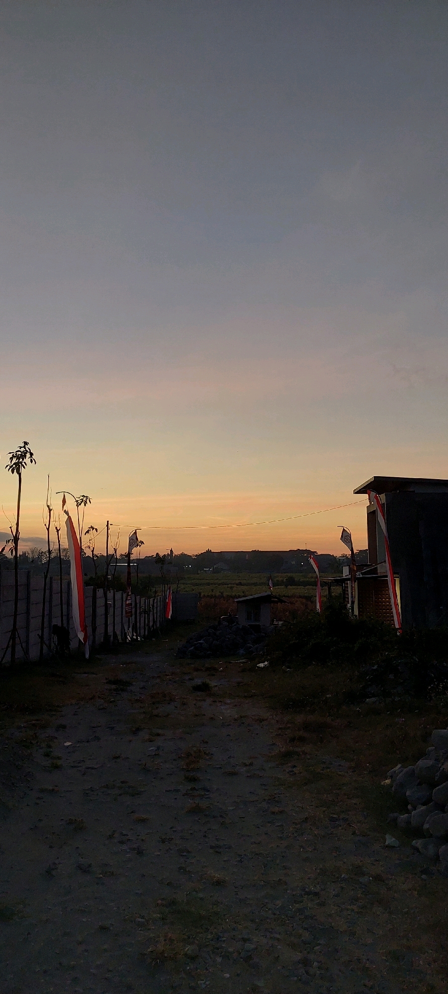 The image depicts a twilight scene with a clear, gradient sky transitioning from a pale grey at the top to warm orange and yellow hues near the horizon. In the foreground, a dirt path leads into the scene, flanked by a grey concrete fence on the left and a dark-colored building on the right. Several Indonesian flags, red and white, are prominently displayed, some attached to poles and others affixed to the fence or building.
Along the left side of the path, the concrete fence is adorned with several Indonesian flags. Bare trees with thin branches are scattered along the fence line. On the right side of the path, a modern-looking dark building with a flat roof is visible. An Indonesian flag is attached to the side of this building, and another is seen flying from a pole in front of it.
The environment appears to be somewhat rural or semi-urban, with hints of distant structures and vegetation. The ground is rough and uneven, covered with dirt and scattered rocks. The lighting suggests it is either dawn or dusk. There are no people visible in the image.